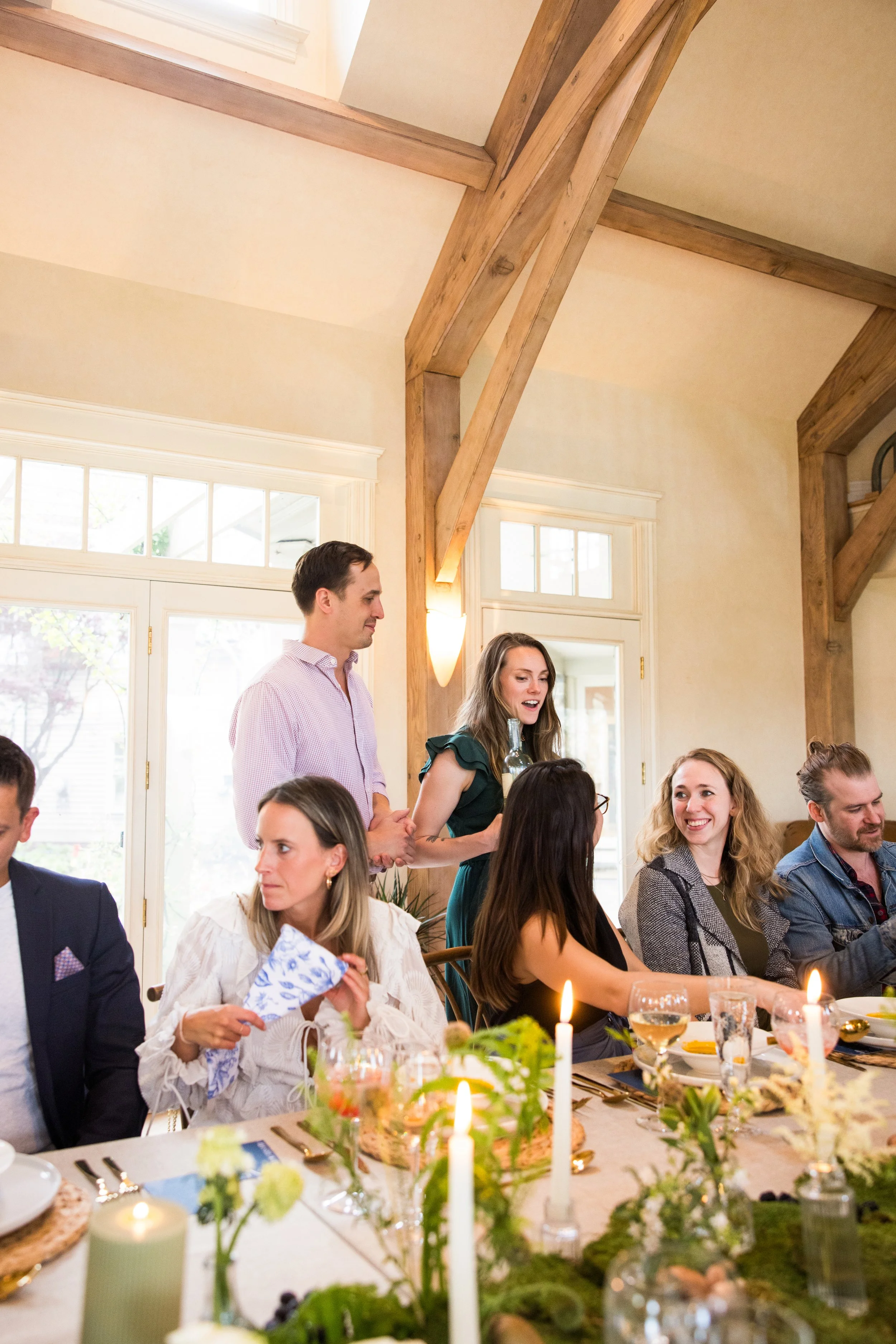 People gathered for a celebration or dinner party, with a woman standing and speaking, holding a bottle, near a long table decorated with candles and flowers in a bright room with wooden beams.
