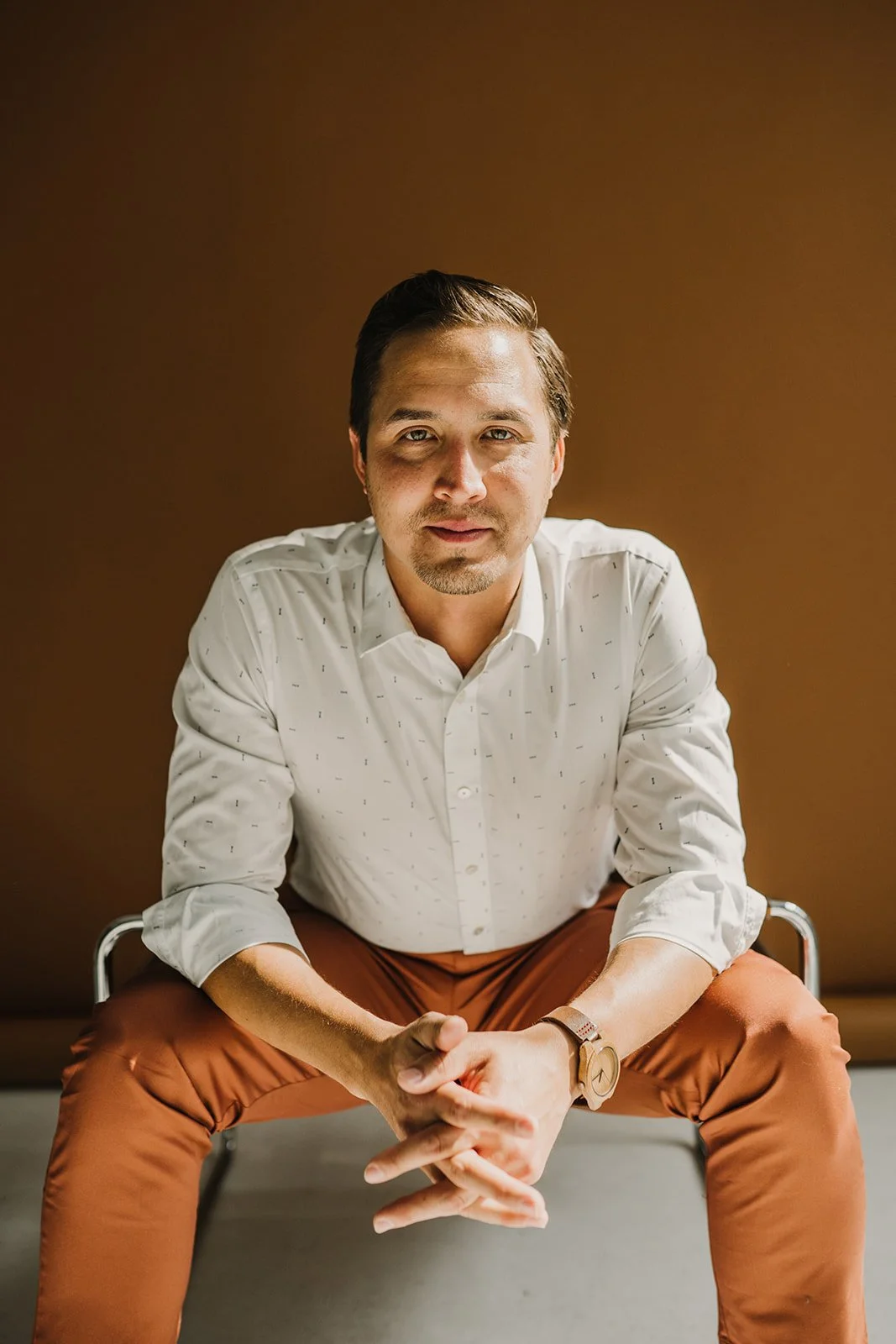 A young man with dark hair, wearing a white button-down shirt and brown pants, sitting on a transparent chair against a brown background, looking directly at the camera.