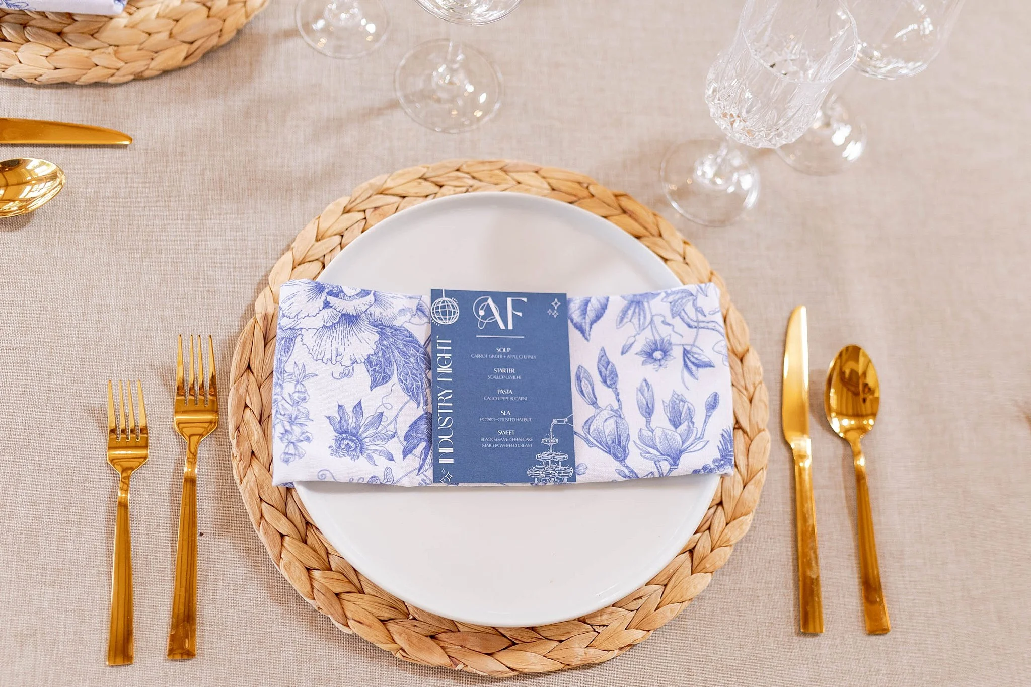 Set table with a white plate on a woven placemat, gold utensils, and a blue floral napkin with a menu. Clear glasses and a water glass are visible on a beige tablecloth.