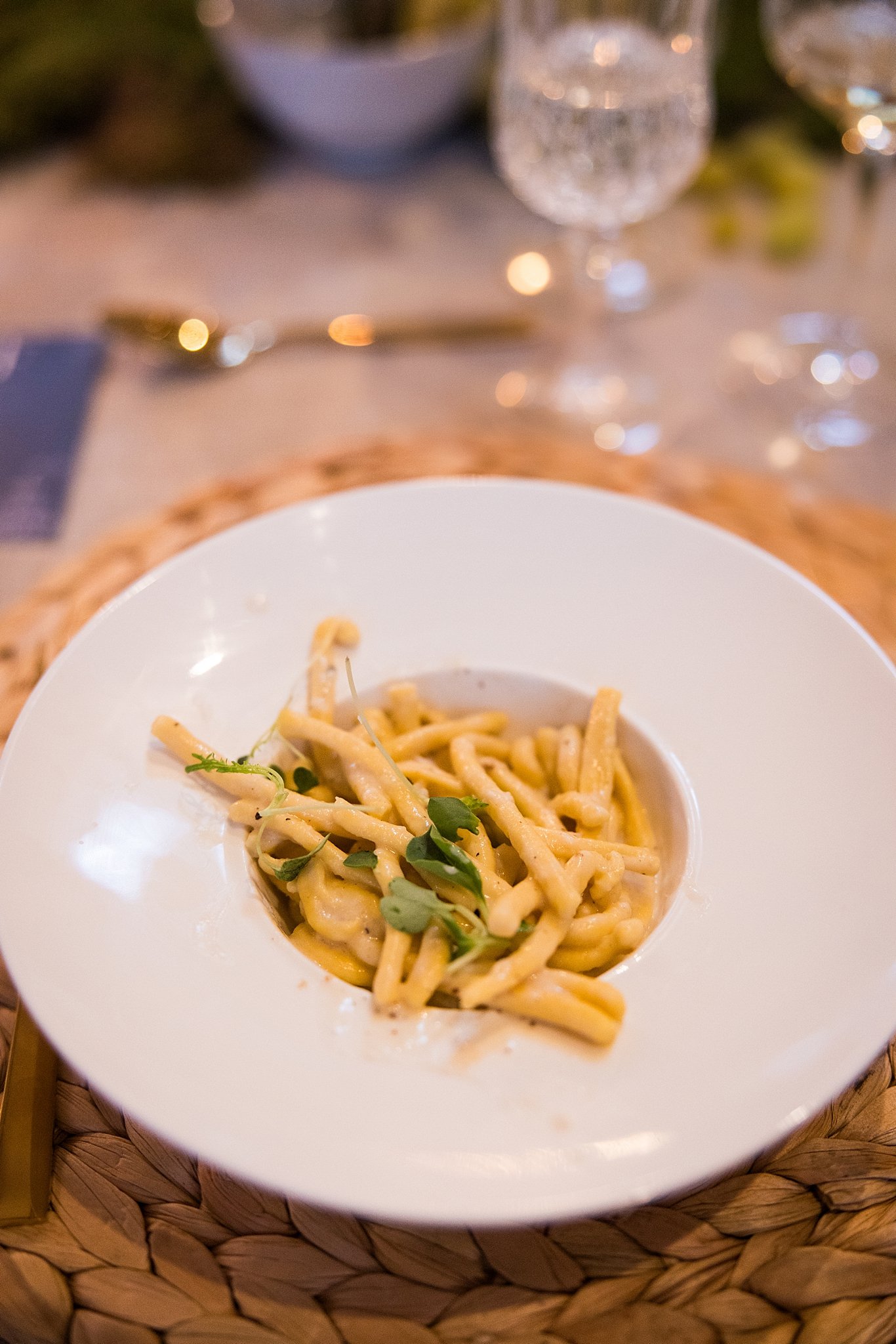 A white plate of creamy pasta garnished with microgreens on a woven placemat, with a blurred background of a table setting and glassware.