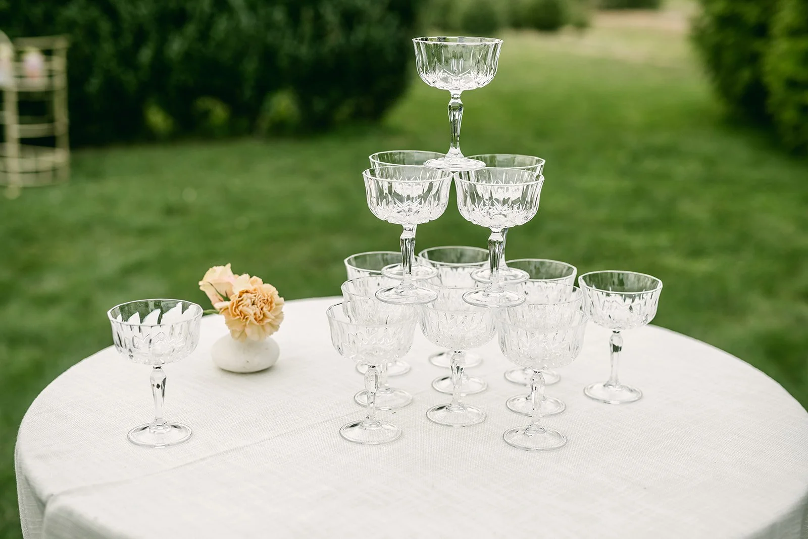 Stacked crystal glasses on a white tablecloth with a small flower arrangement in a white vase outdoors.