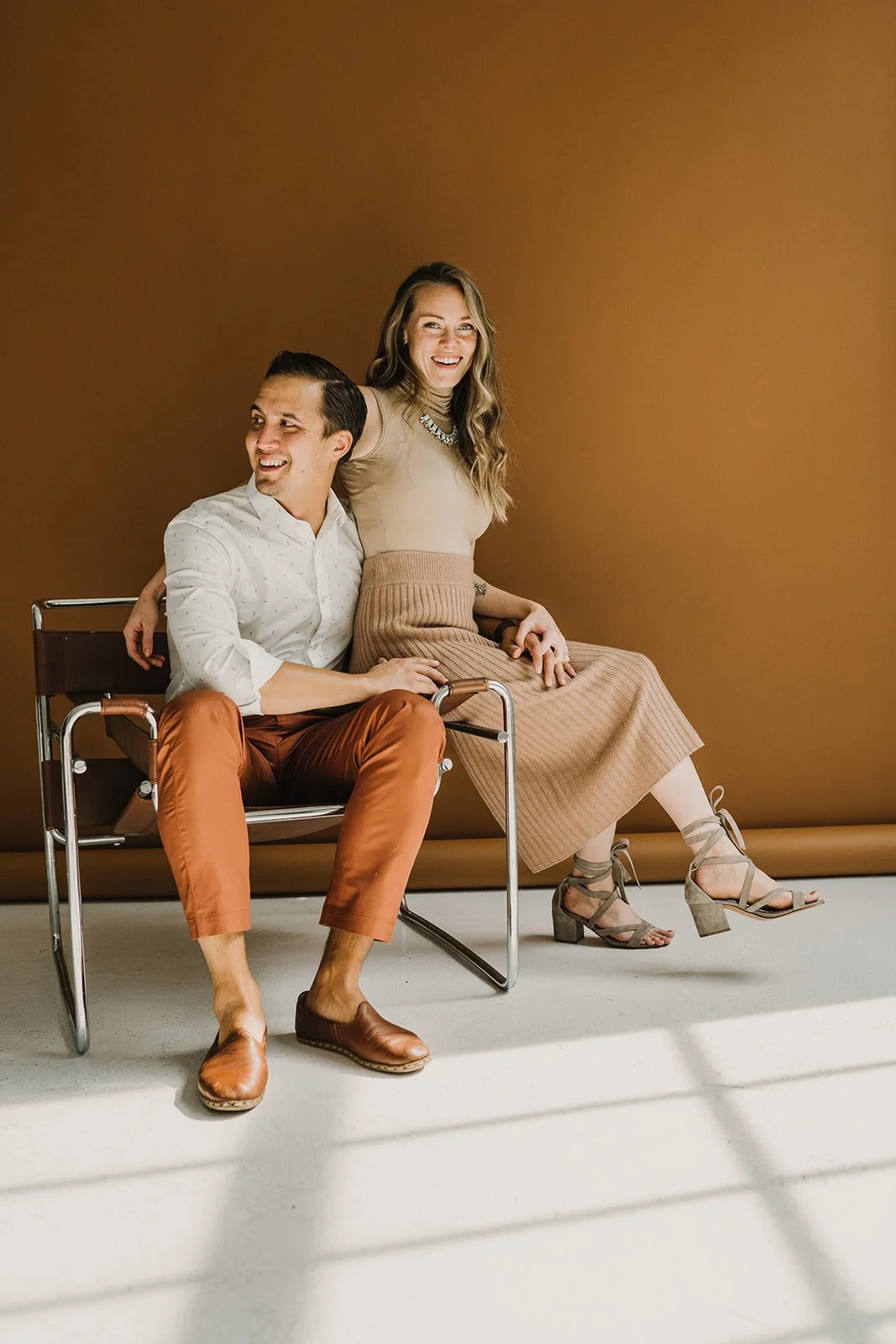 A man sitting on a chair and a woman sitting on his lap, both smiling and looking away from the camera, against a brown background.
