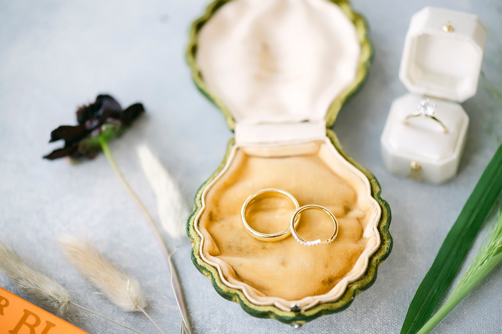 Two wedding rings inside a vintage green and cream seashell-shaped jewelry box, surrounded by dried flowers, green grass, and a book.