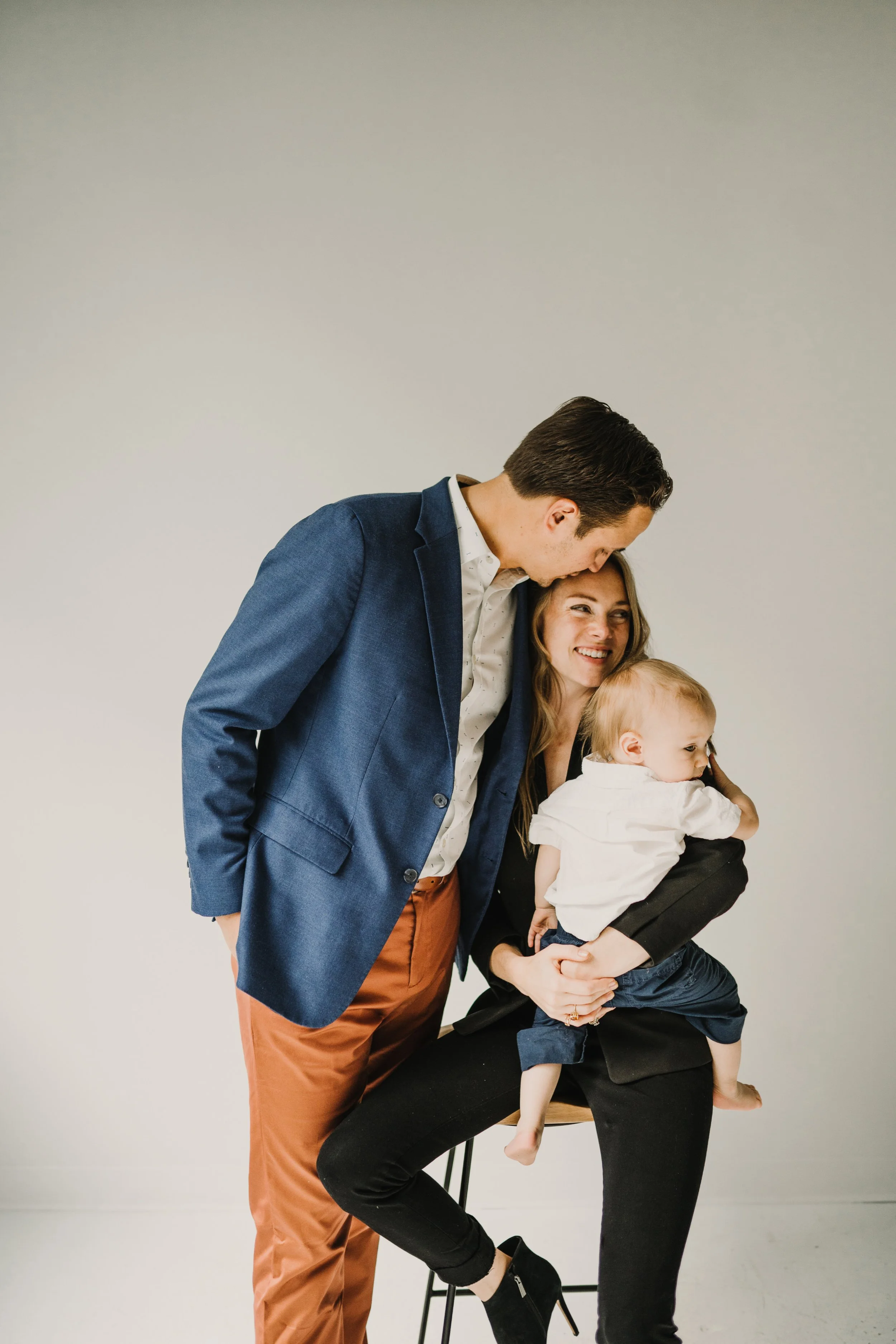 A family of three enjoying a moment together. The man in a blue blazer and brown pants leans toward the woman. The woman, sitting on a chair, is holding a young child, and all are smiling and sharing a happy moment.