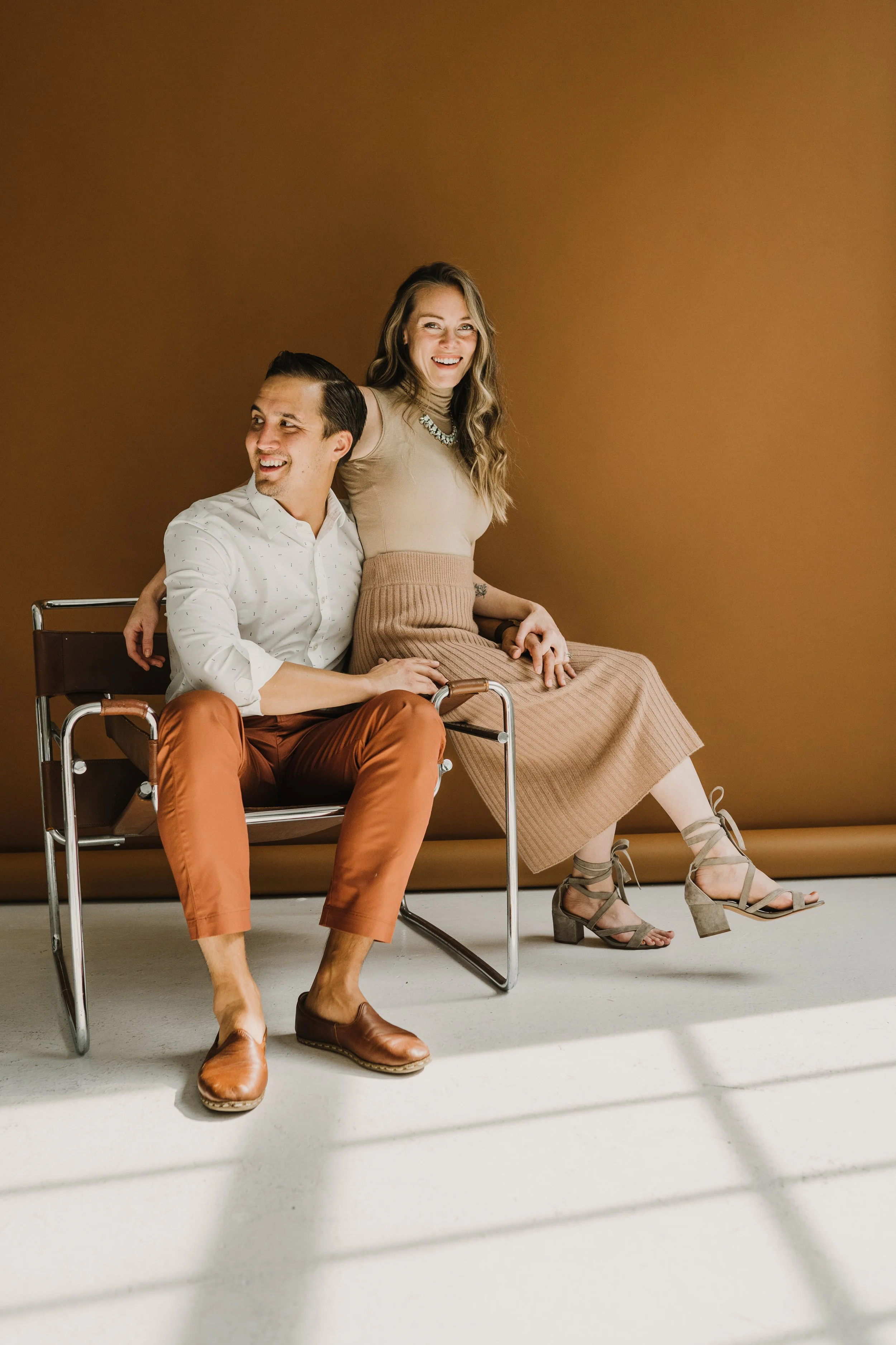 A man sitting on a chair with a woman sitting on his lap, both smiling and laughing in front of a brown background.