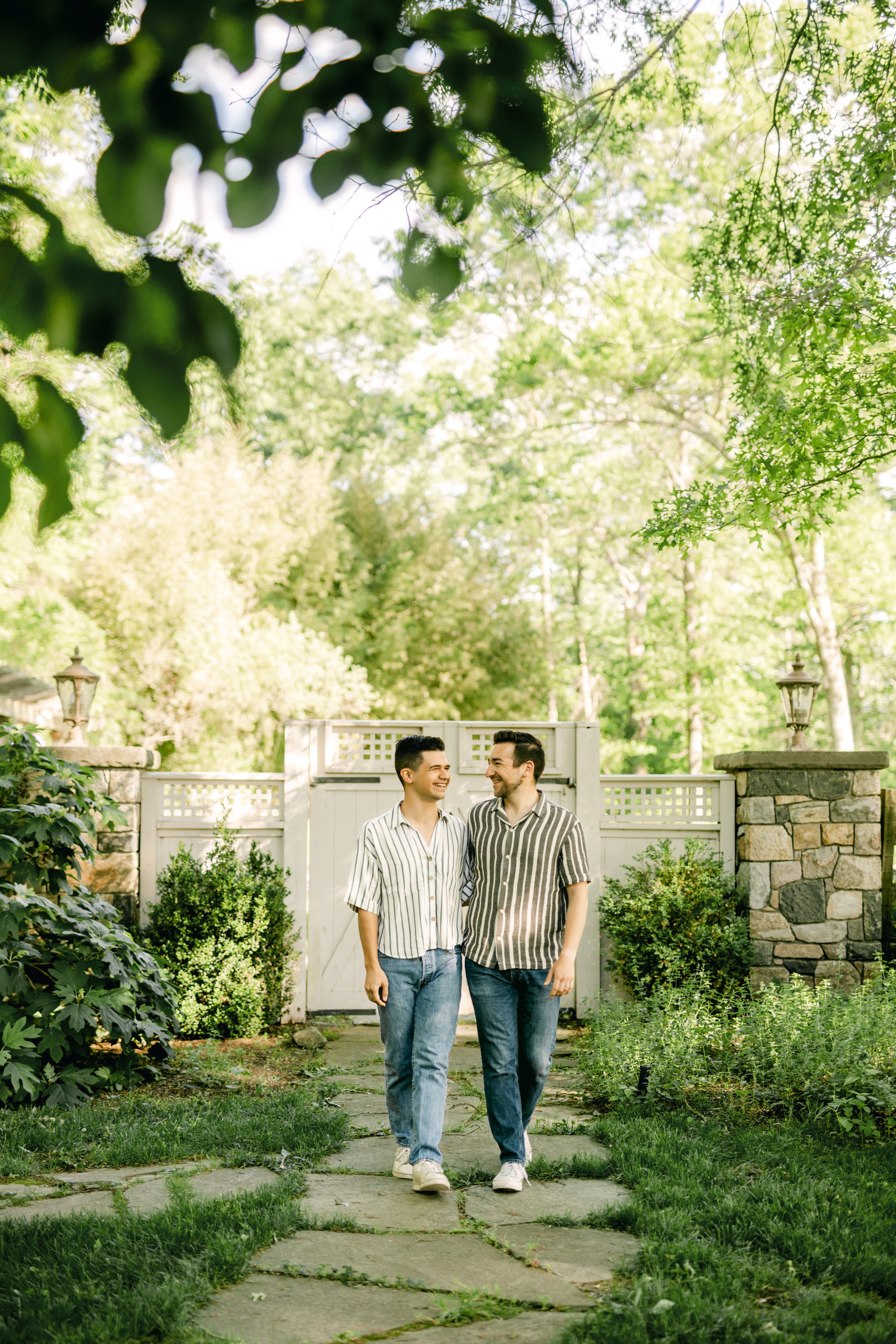 Two men smiling and walking together in a garden with lush greenery and a stone pathway, a white fence, and stone pillars with lanterns in the background.