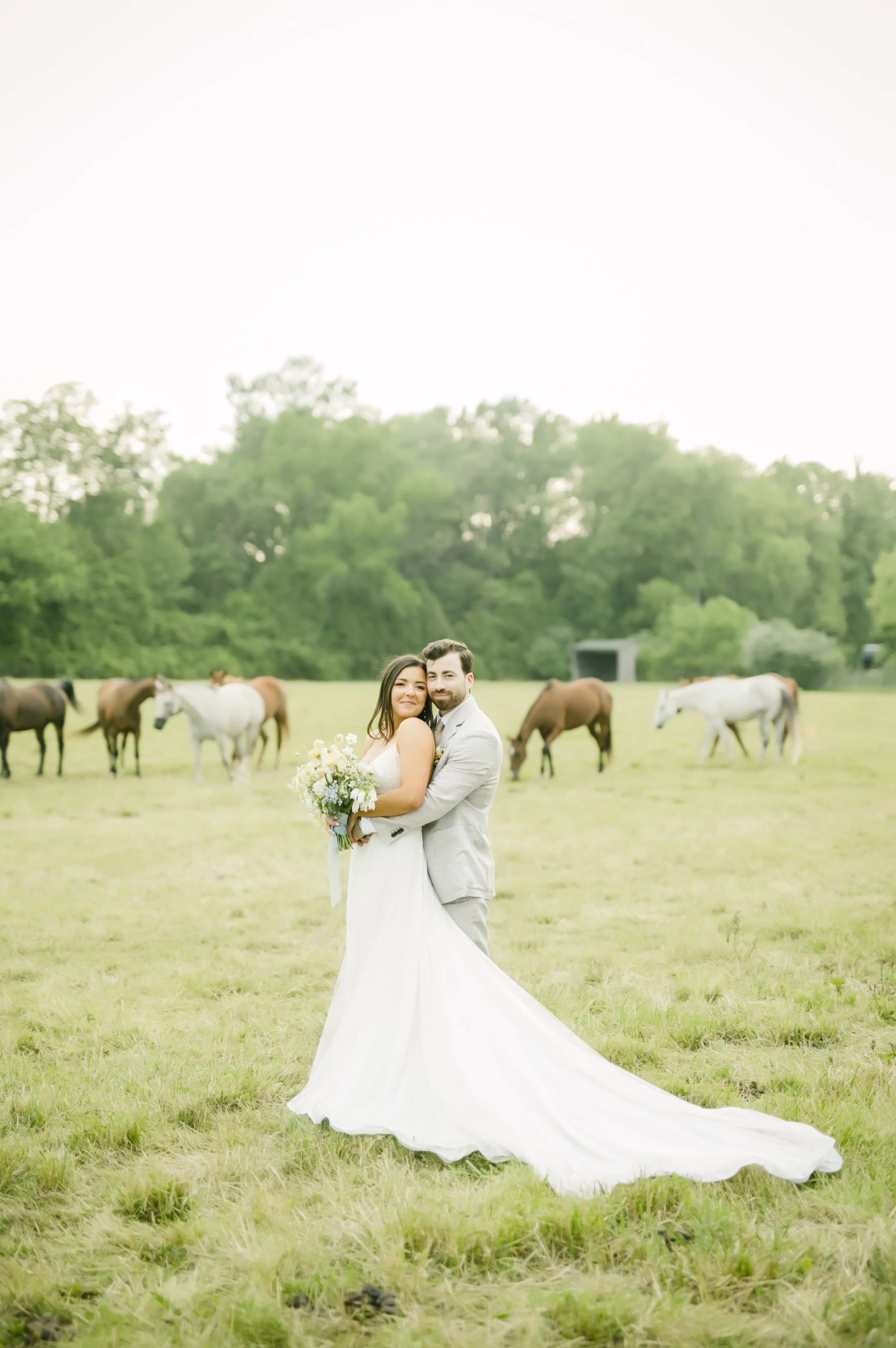 A newlywed couple in wedding attire standing on a grassy field with a group of horses in the background, lush green trees, and a small shed.
