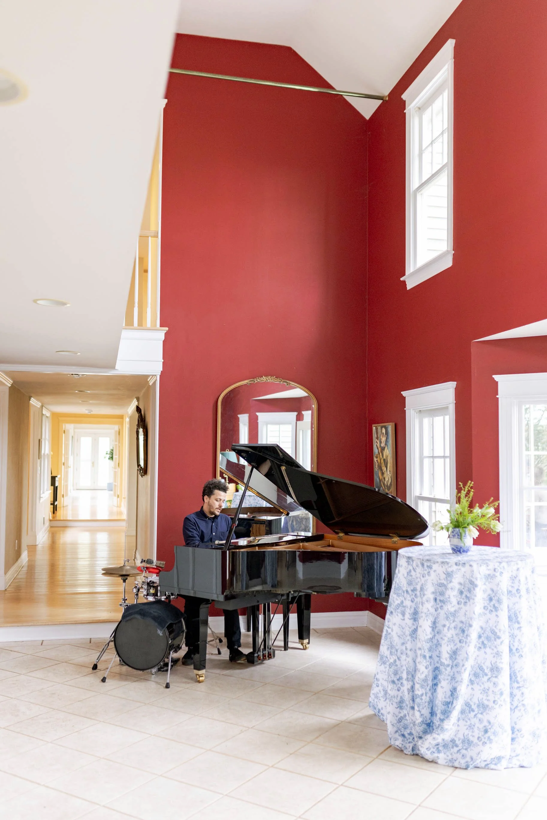 A man playing a black grand piano indoors with red walls, large windows, a mirror, and a table with a floral tablecloth and a flower arrangement.