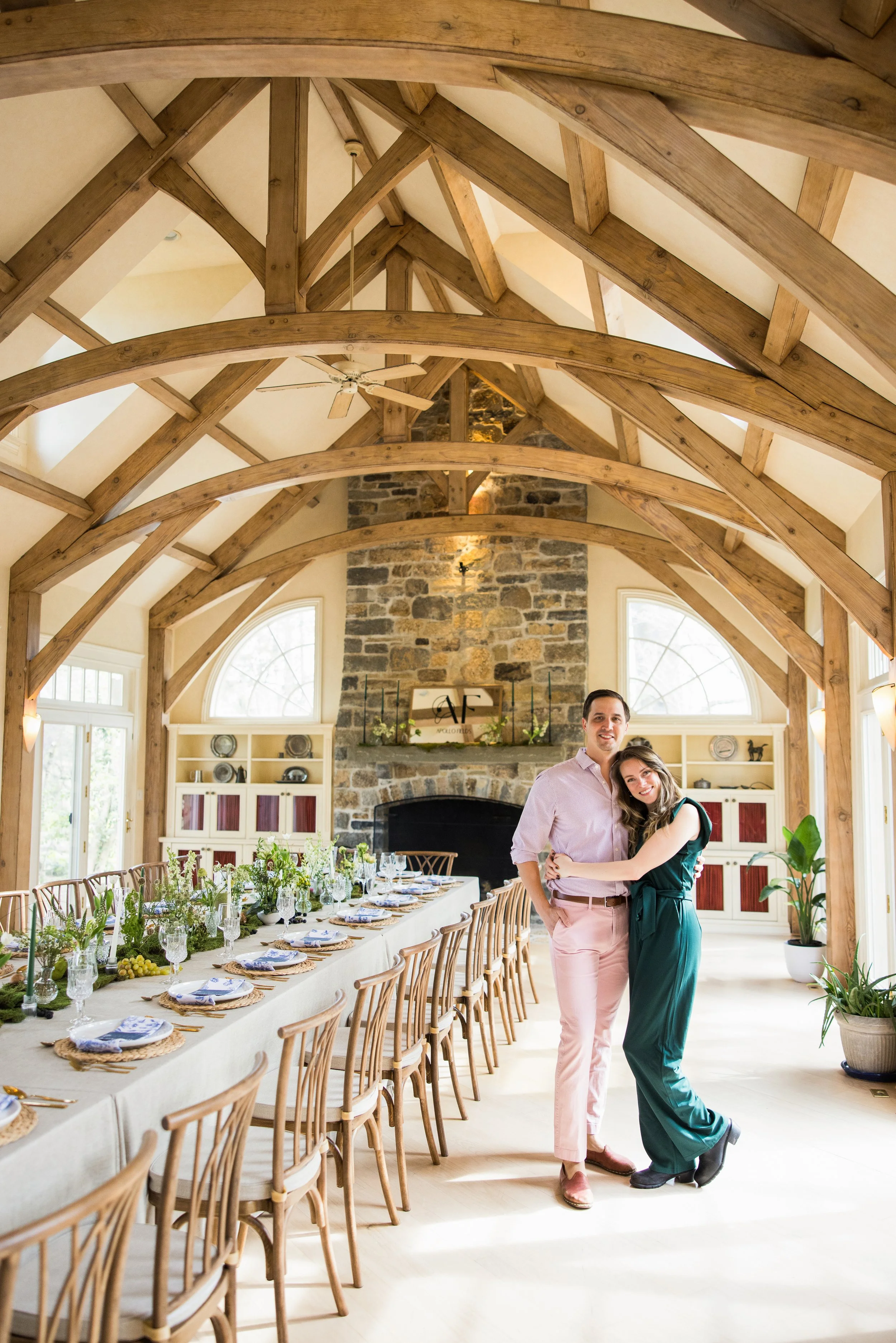 A couple standing together inside a spacious dining area with a high, vaulted wood ceiling and large windows. The long dining table is decorated with greenery and place settings, and a stone fireplace is visible in the background.