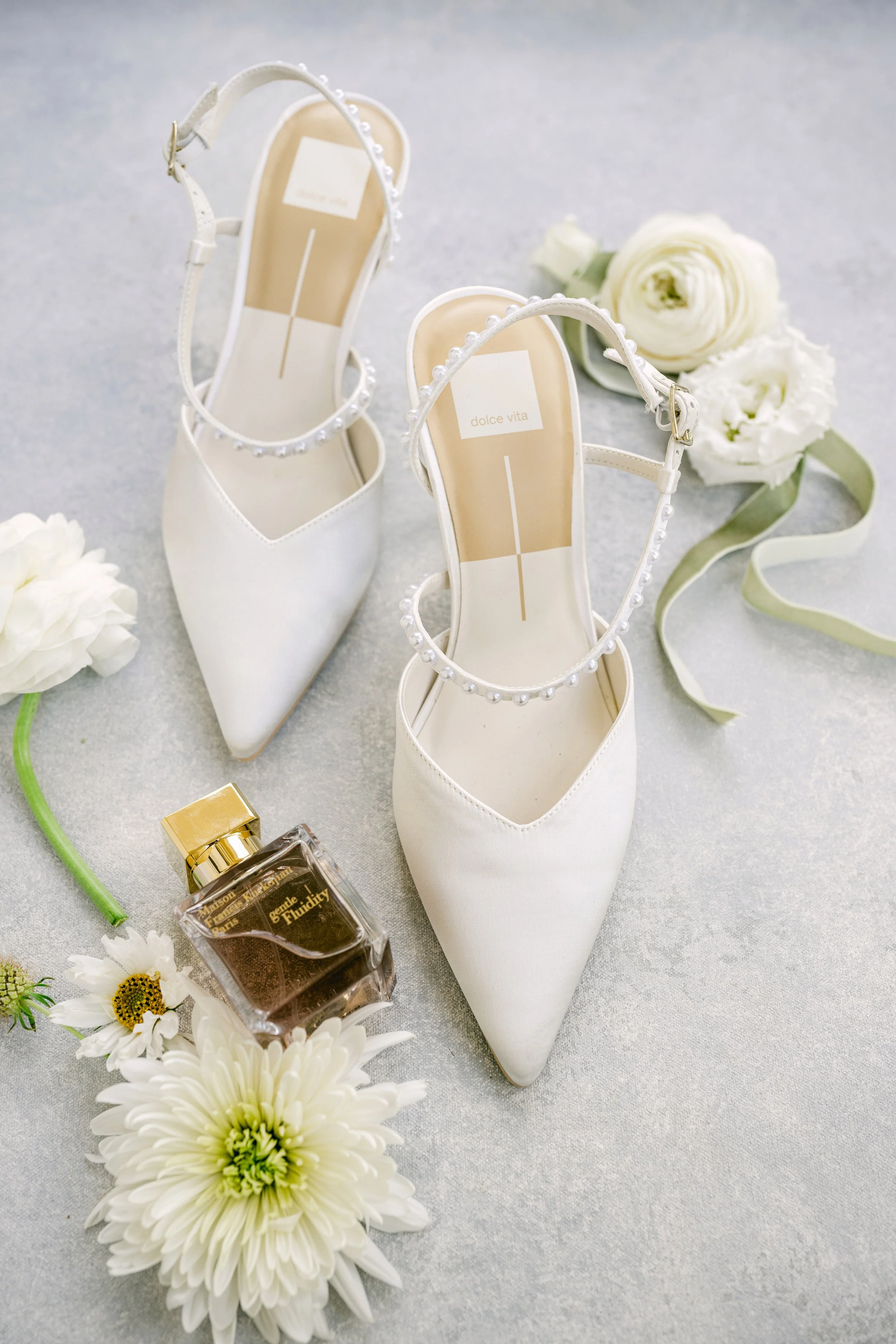 White high-heeled shoes with ankle straps decorated with pearls, a bottle of perfume, white flowers, and a soft grey surface.