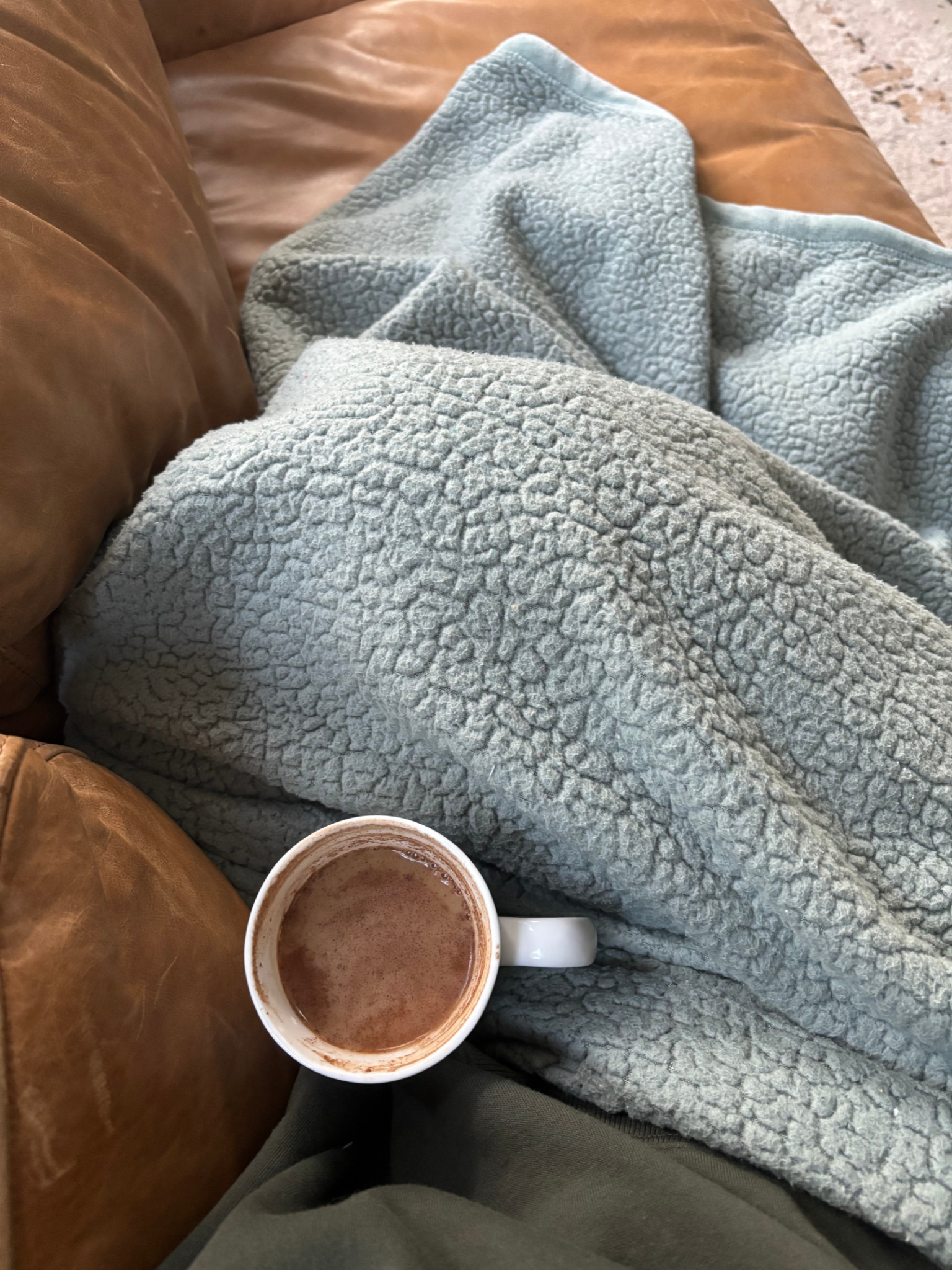 Close-up of a person's lap covered with a textured, light gray fleece blanket, a brown leather couch armrest on the left, and a white mug filled with hot chocolate in the foreground.