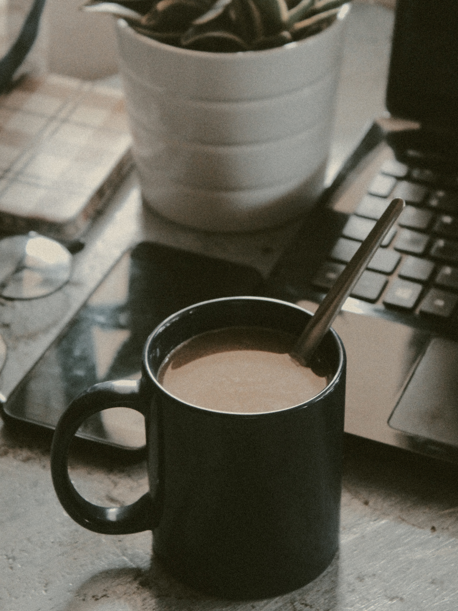 A black coffee mug filled with coffee, placed on a table near a laptop and a potted plant.