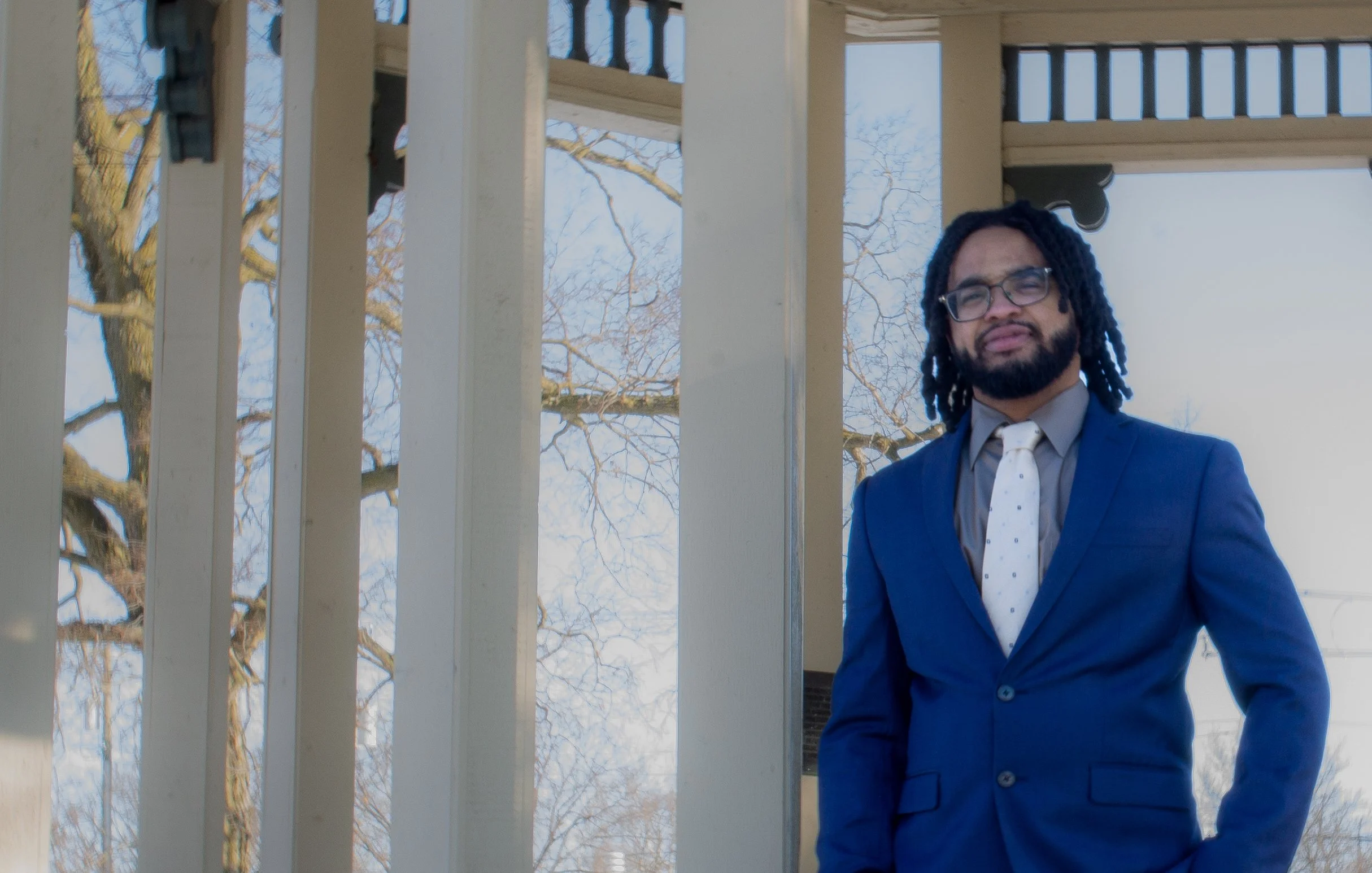 A man in a blue suit and white tie standing outdoors near a porch with white railings, with trees and a blue sky in the background.