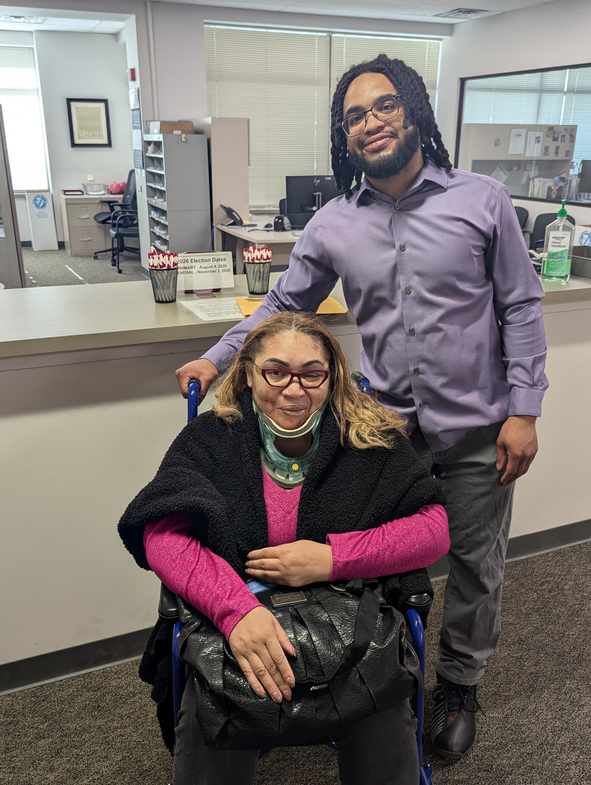 A man in a purple shirt and glasses standing behind a seated woman in a wheelchair, in an office reception area. The woman is wearing glasses, a pink shirt, and a black jacket, with a neck brace and a face mask hanging below her chin. There are donation jars, a sign with election dates, and office supplies on the countertop.