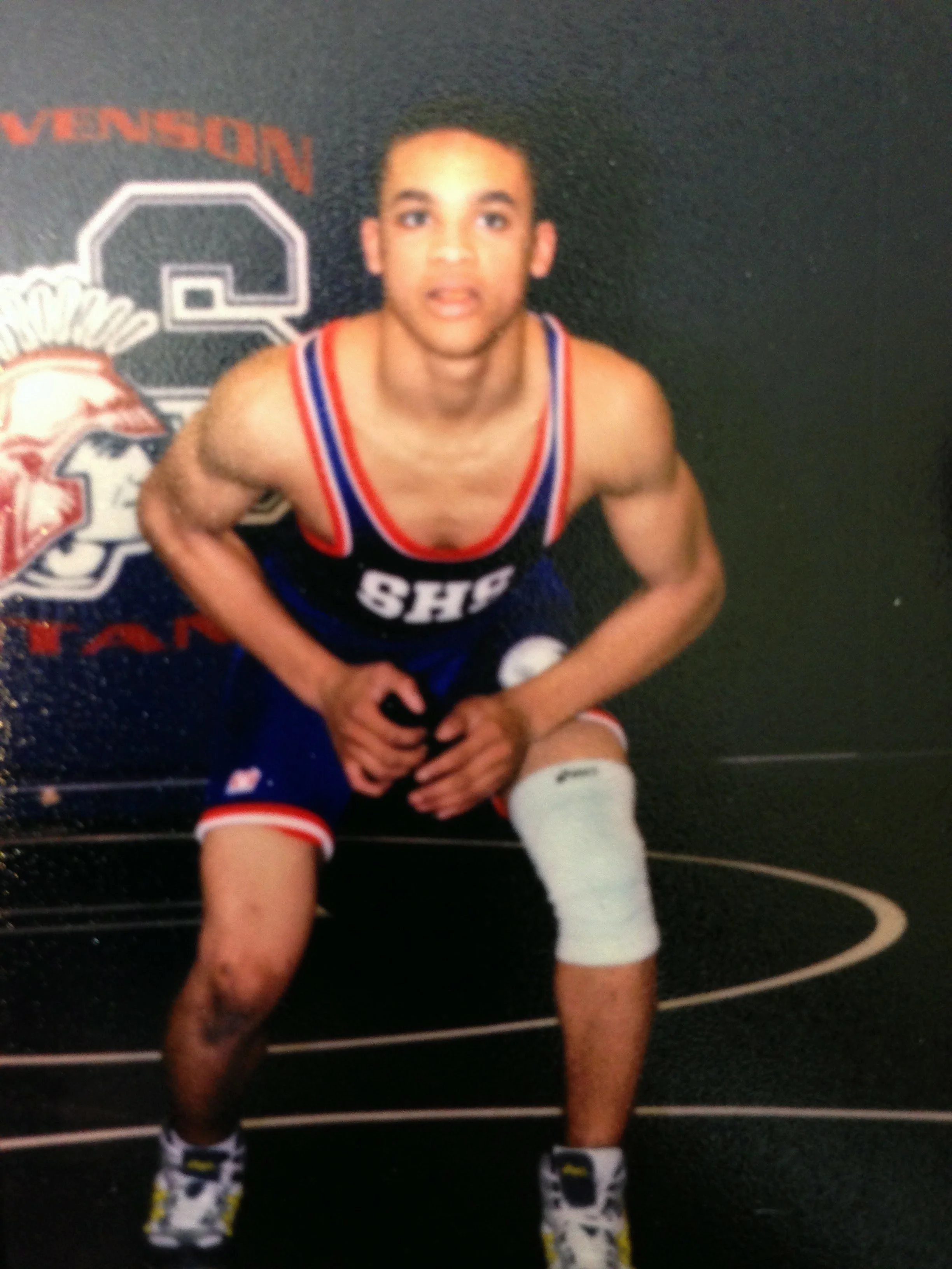 Young male wrestler in athletic gear crouching in a wrestling stance, with a knee brace on his right leg, on a wrestling mat with a sports-themed backdrop.