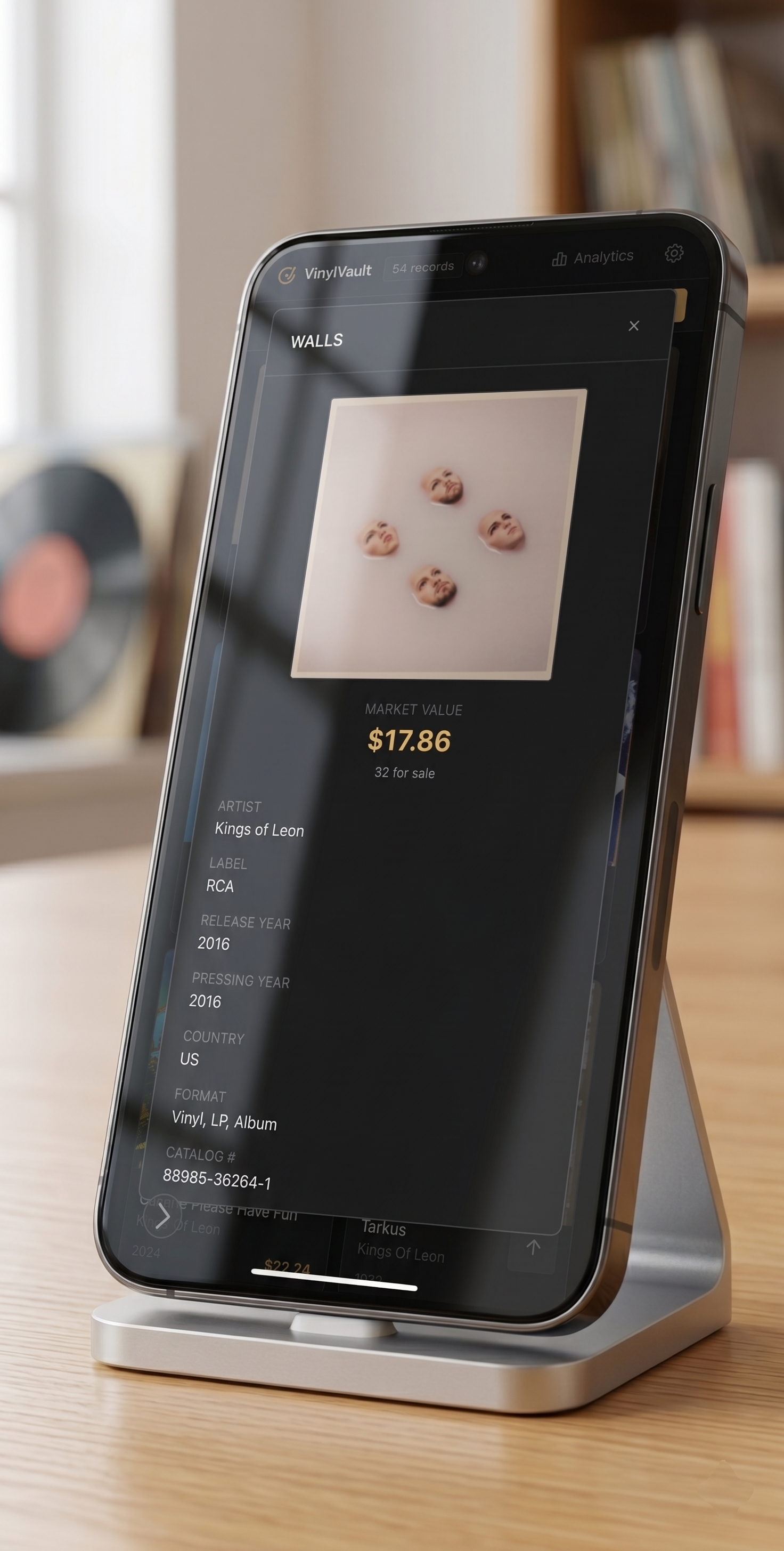 A smartphone displays information about a record titled "Kings of Leon" by RCA, released in 2016, with a retail value of $17.86, on a wooden table in a well-lit room.
