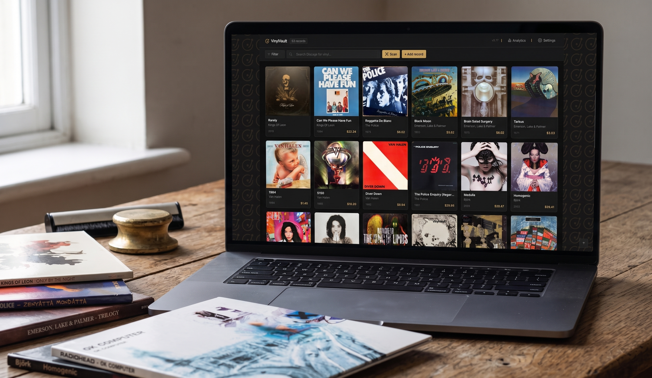 A laptop on a wooden desk displaying a music streaming app with album covers, surrounded by magazines and books.