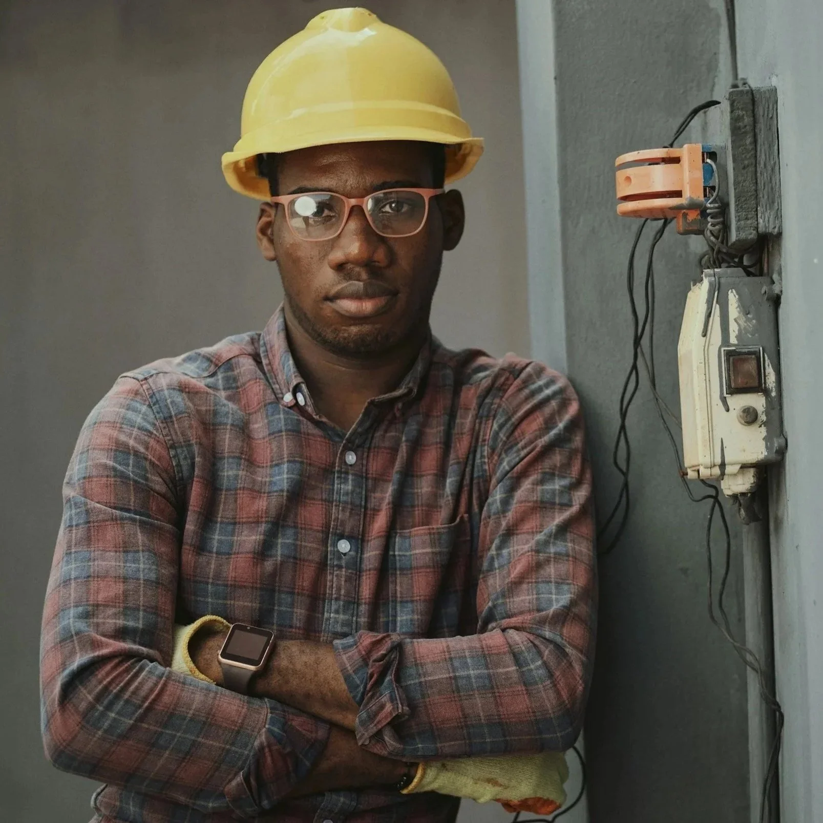 A man wearing glasses, a yellow construction helmet, a plaid shirt, and a smartwatch stands with arms crossed near an electrical outlet on the wall.