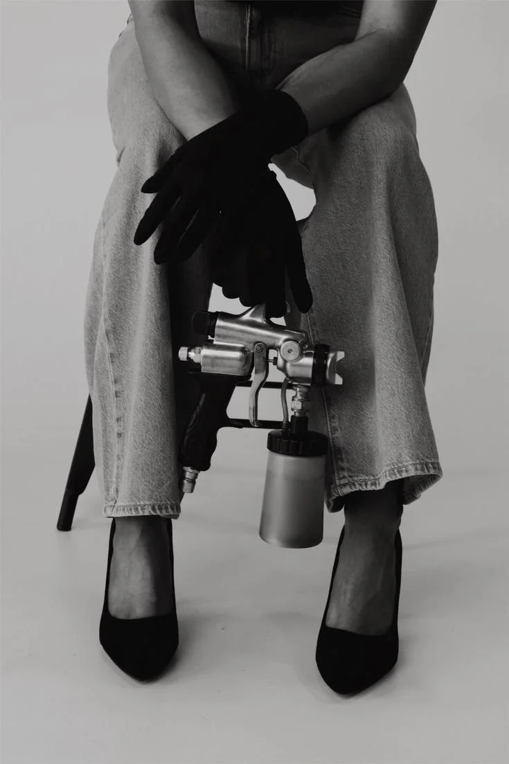 Person sitting with crossed hands holding a silver spray tan gun while wearing black gloves, light jeans, and black high heels.