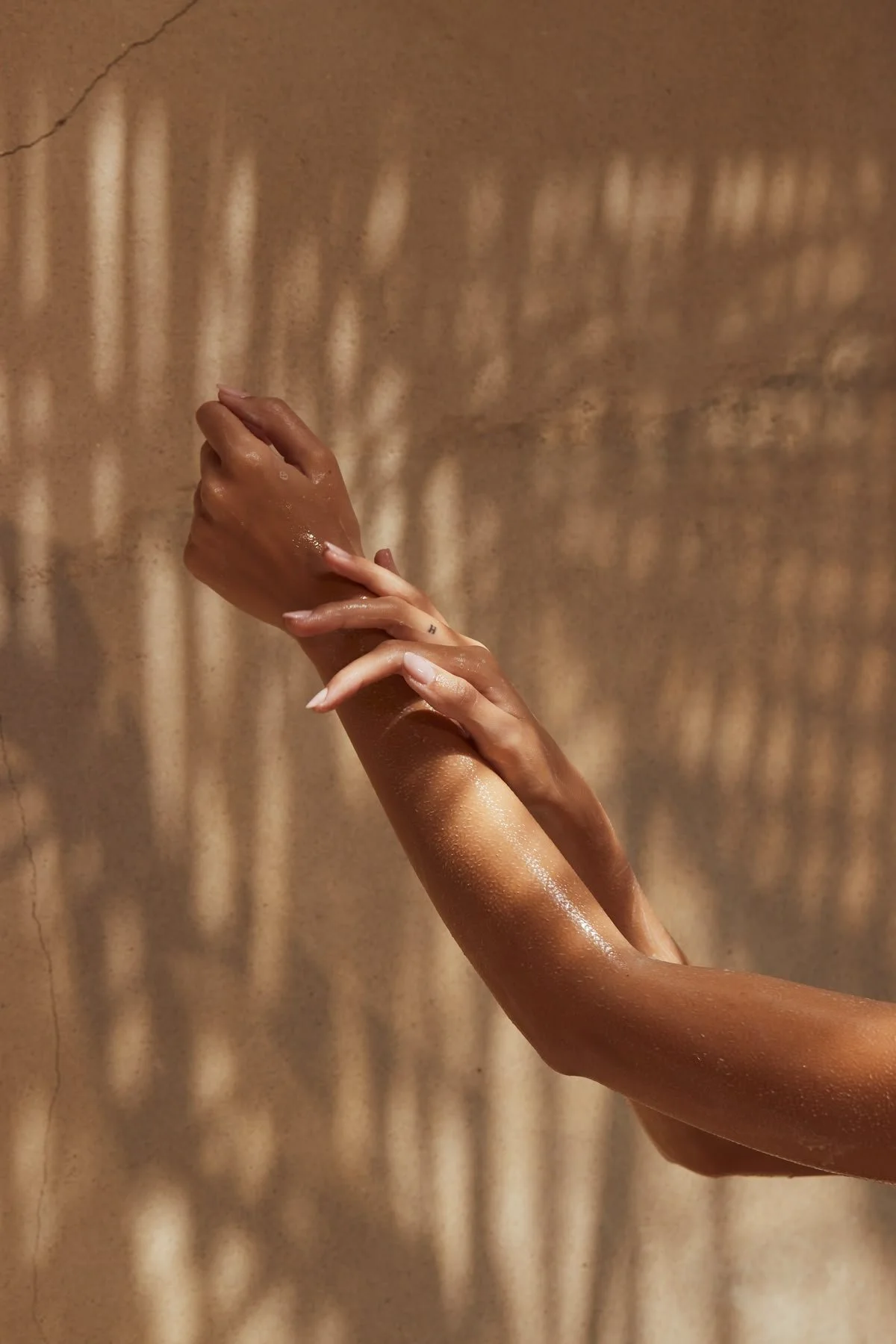 Close-up of two hands with a warm brown background and shadow patterns, one hand gently touching the wrist of the other.