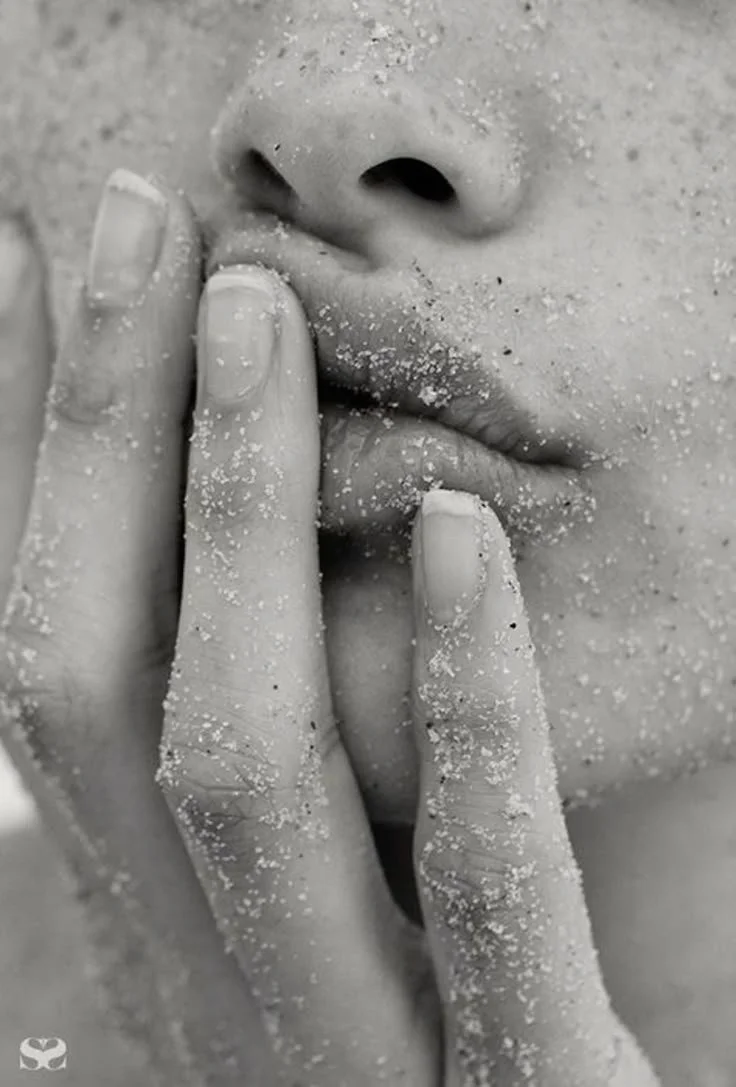 Close-up of a person's lips and fingers covered in sand, with fingers touching their lips.