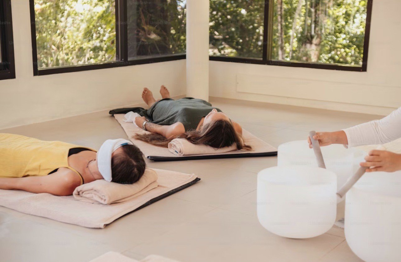 Two women lying on mats with heads on pillows, receiving sound healing using crystal bowls in a room with large windows and greenery outside.
