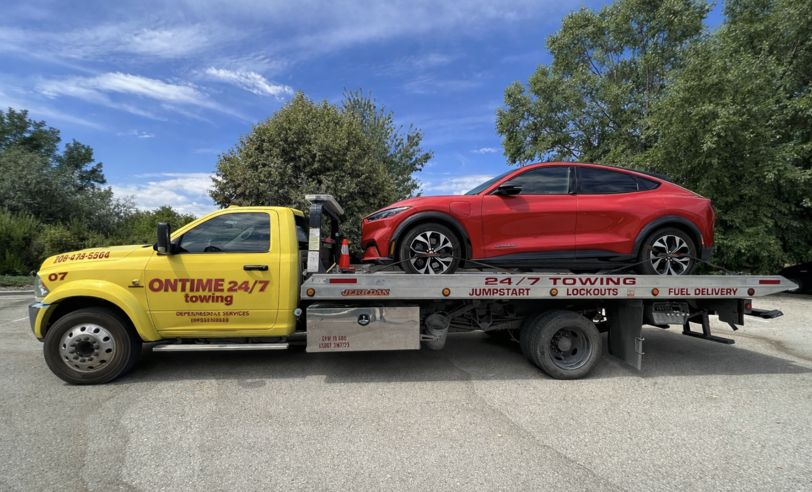 Red car loaded on a yellow tow truck with green trees and a blue sky with clouds in the background.