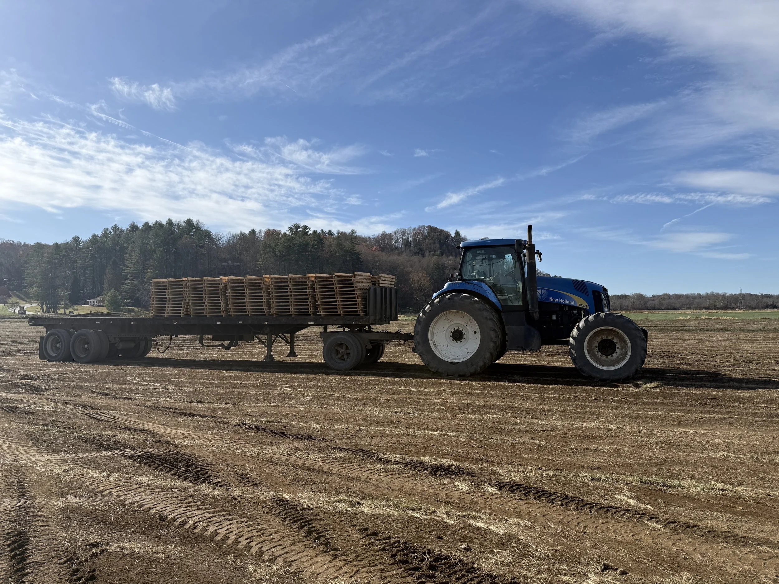 A blue tractor pulling a flatbed trailer with stacked pallets on a freshly plowed dirt field under a partly cloudy sky.