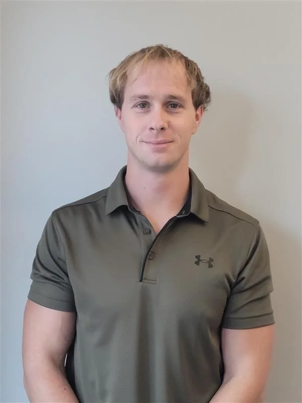A young man with light brown hair and fair skin, wearing a dark green polo shirt with the Under Armour logo, standing against a plain light gray wall.