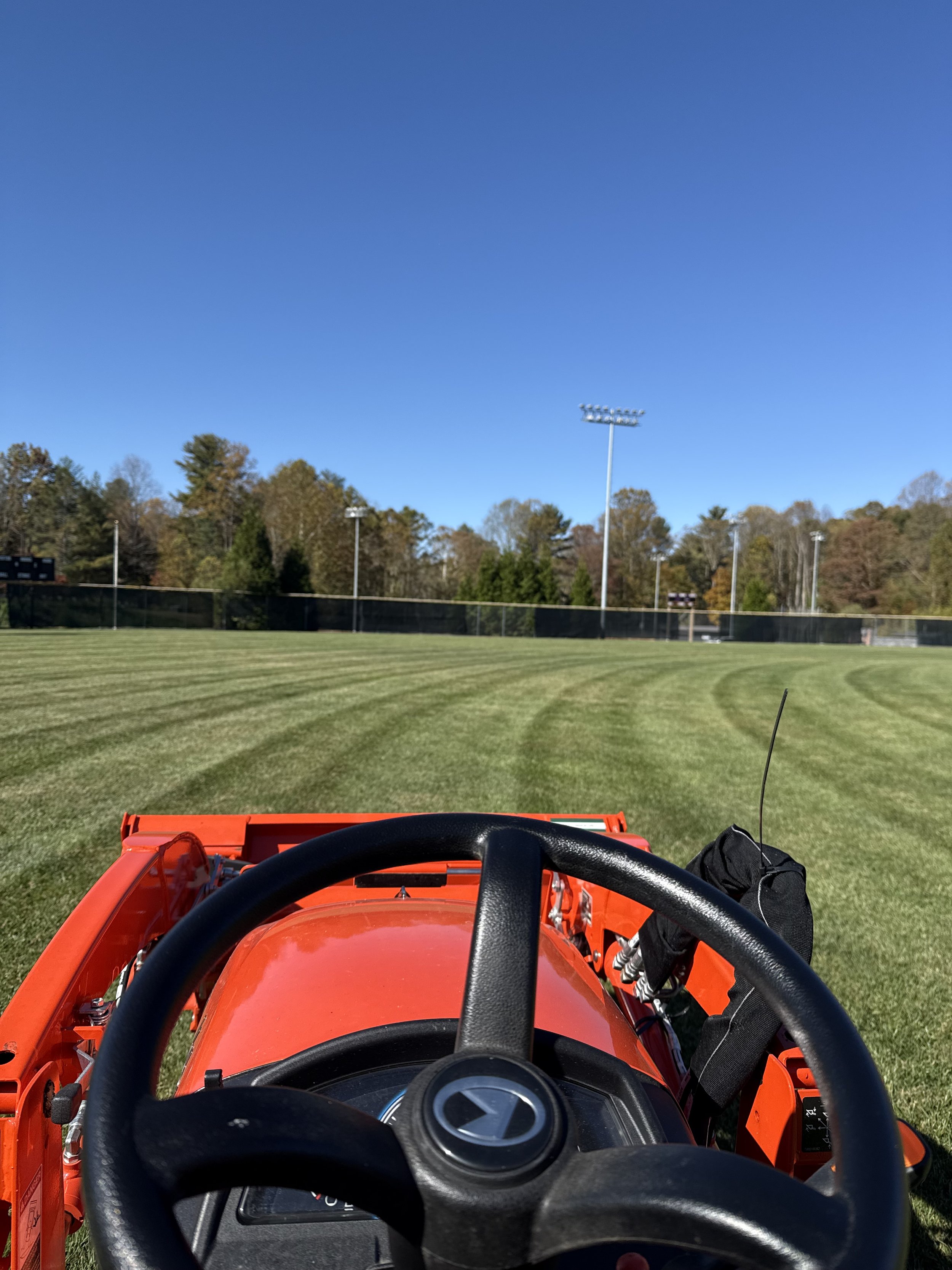 Front view of a red tractor on a grassy sports field, with a clear blue sky and trees in the background.
