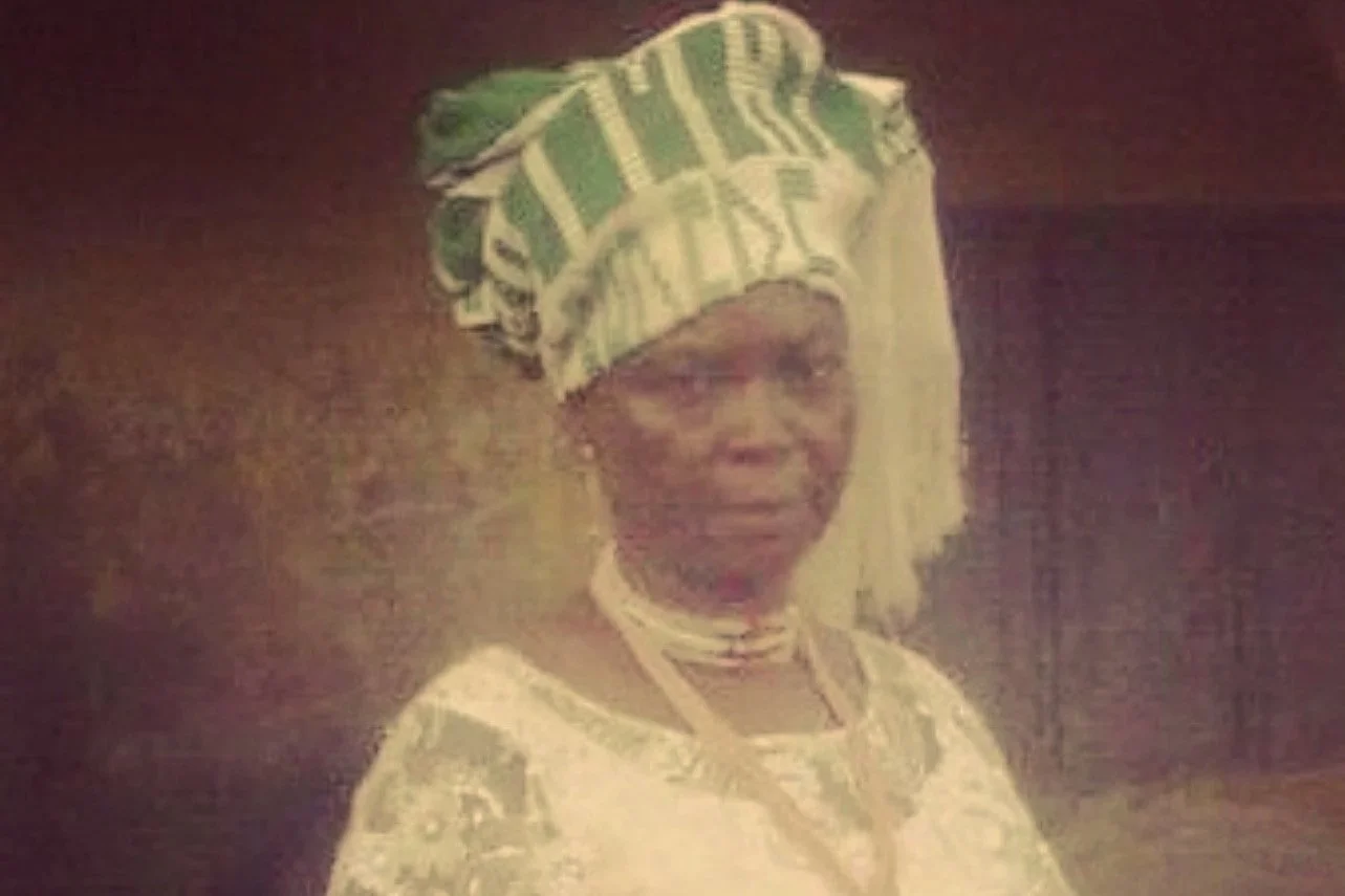 Mulher idosa vestida com roupas tradicionais, usando um leque na cabeça, em um ambiente interno.