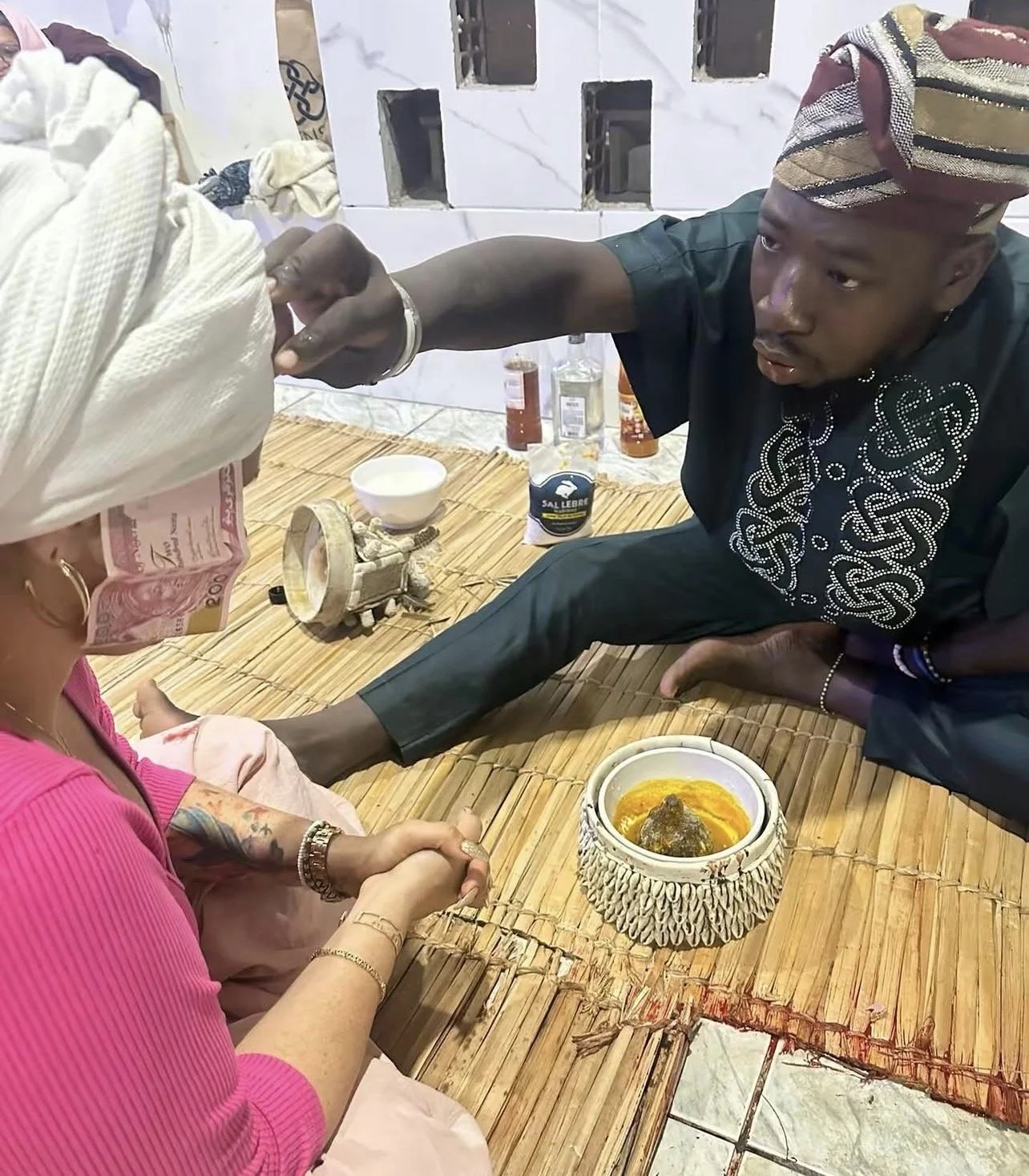 Homem vestindo roupas tradicionais faz oferenda a uma mulher que usa cabeça envolta em um lenço, em um ambiente com teto de alvenaria e objetos tradicionais na mesa.