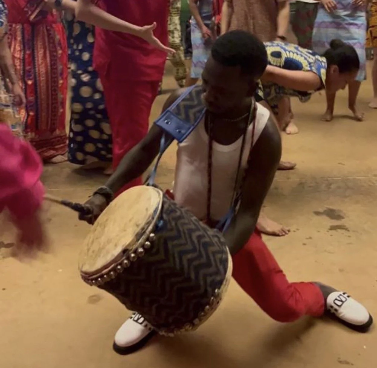Homem tocando tambor durante uma cerimônia ou dança tradicional, cercado por pessoas vestidas com roupas coloridas.