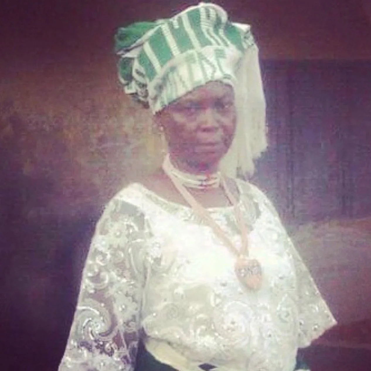 Mulher usando trajes tradicionais com uma cabeça envolta em tecido verde e branco, vestindo roupas brancas com detalhes brilhantes, colar e pulseira, diante de um fundo escuro.