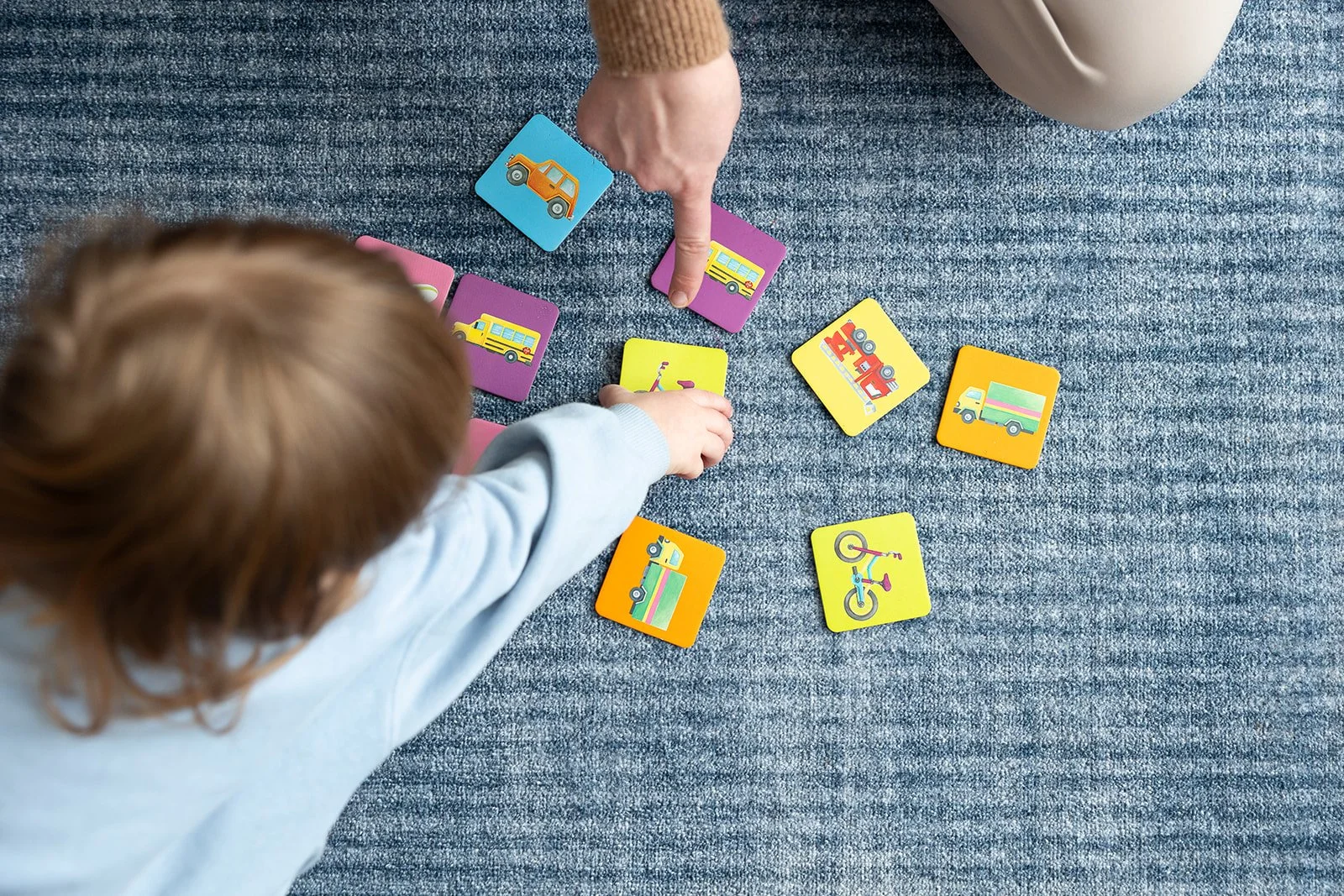 Child and adult playing with colorful illustrated vehicle and transportation-themed flashcards on a carpet.