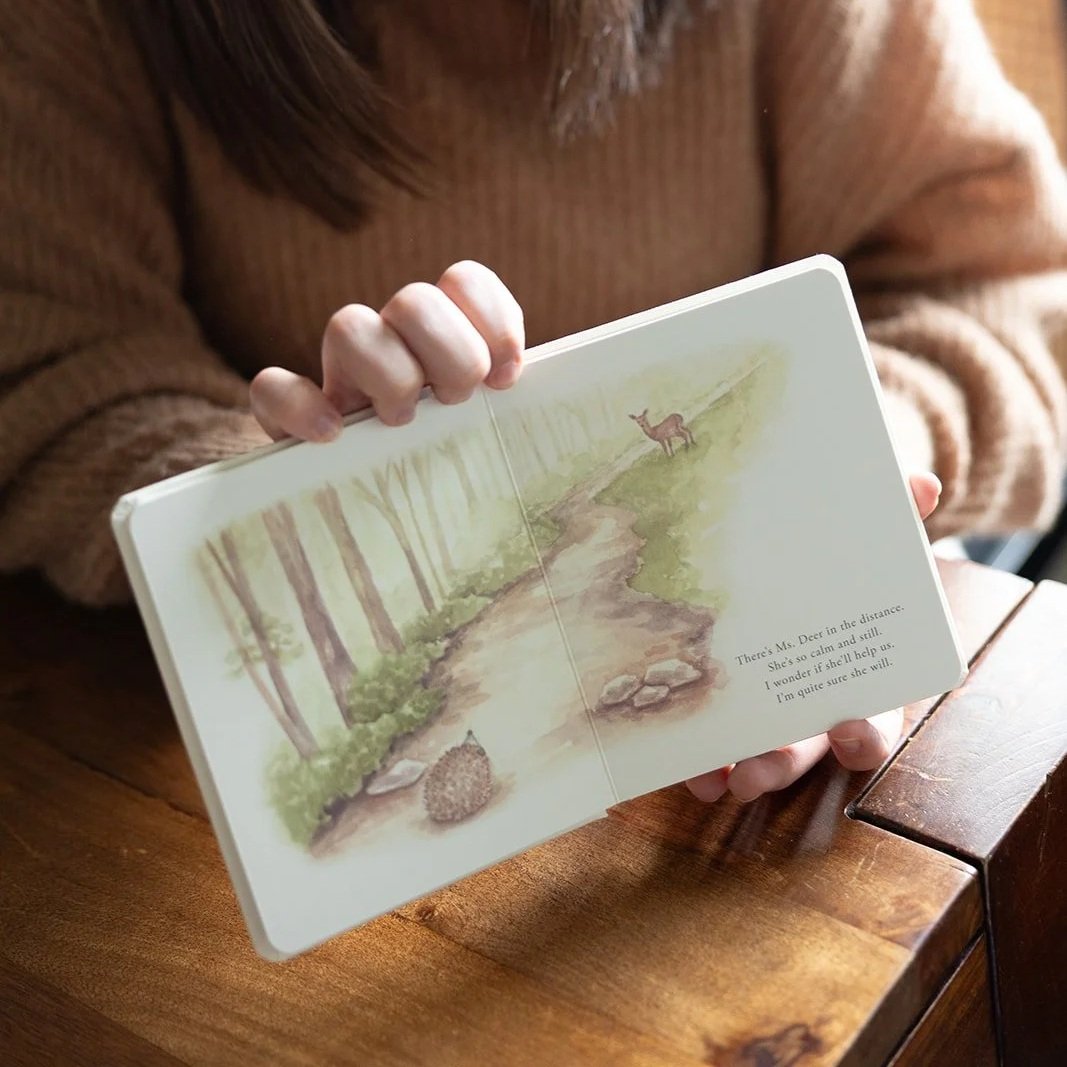 A person holding an open illustrated children's book with a forest and a deer on a trail, with text on the right side.