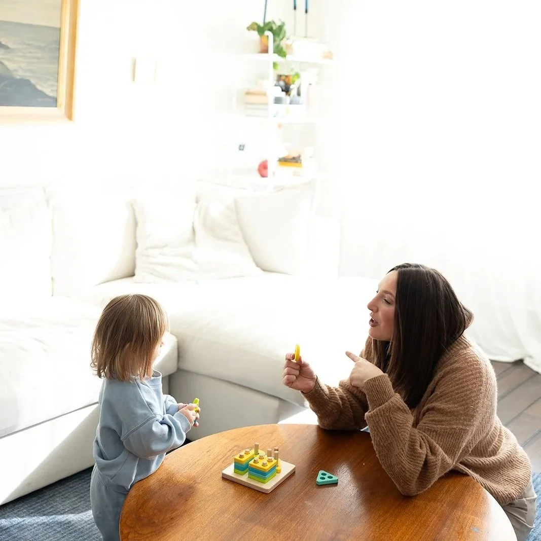 A woman and a young girl playing with colorful stacking toys at a wooden table in a bright living room.