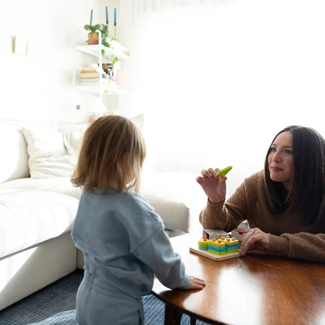 A woman and a young girl playing a board game at a wooden table in a bright living room with white walls and shelves with books and plants.