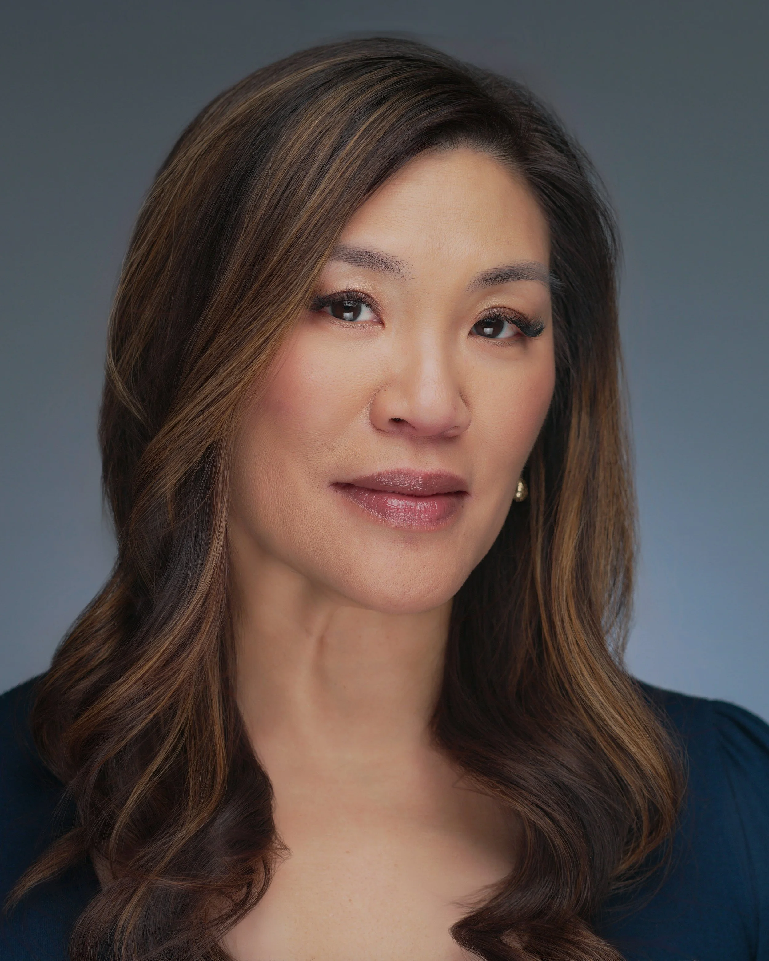 Professional headshot of a woman with shoulder-length brown hair, wearing a dark top, against a gray background.