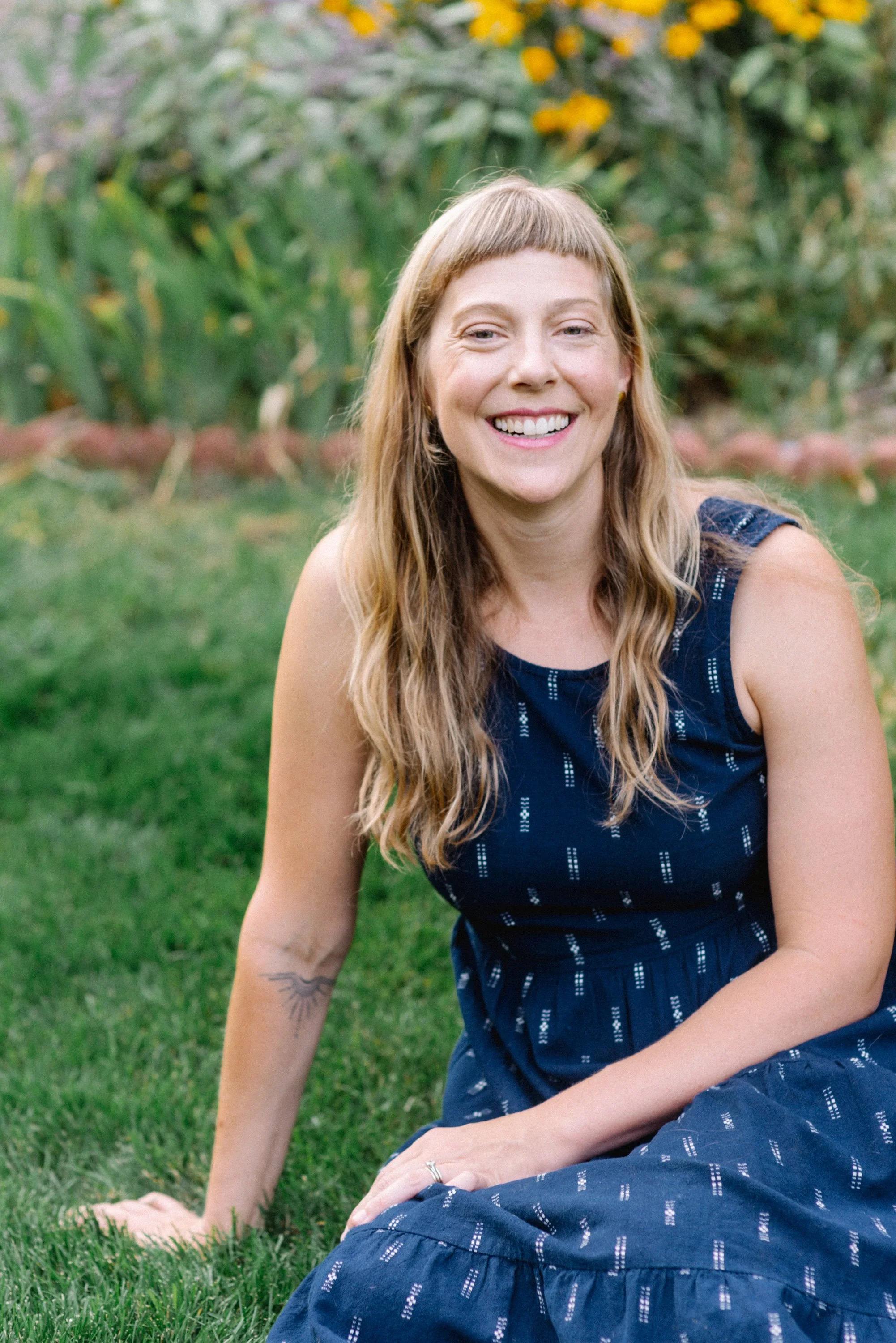A woman with long wavy hair sitting on the grass, smiling, with a garden background of yellow flowers and green plants.