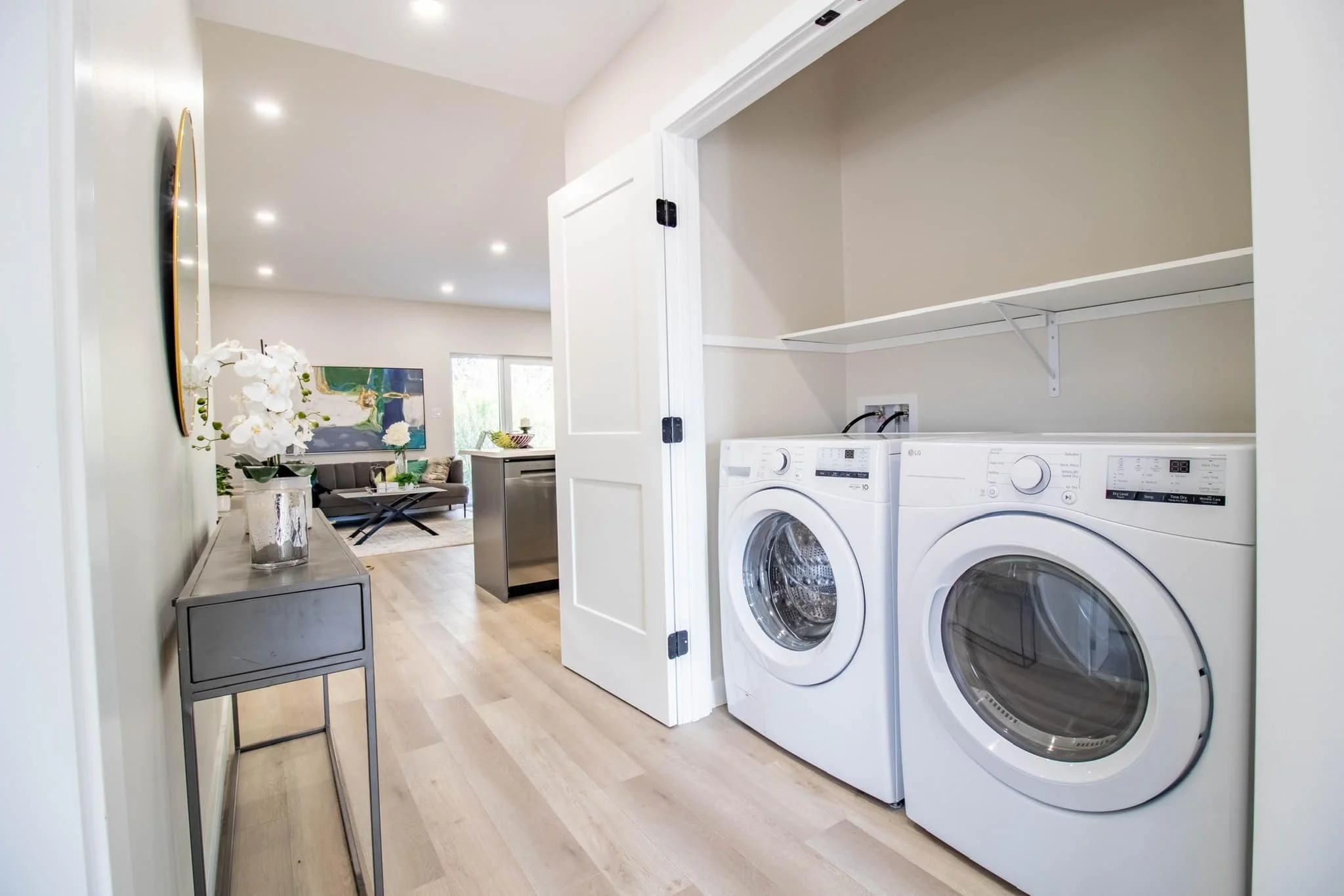 Laundry area with a washer and dryer inside a small closet adjacent to a living room with a sofa and artwork on the wall.