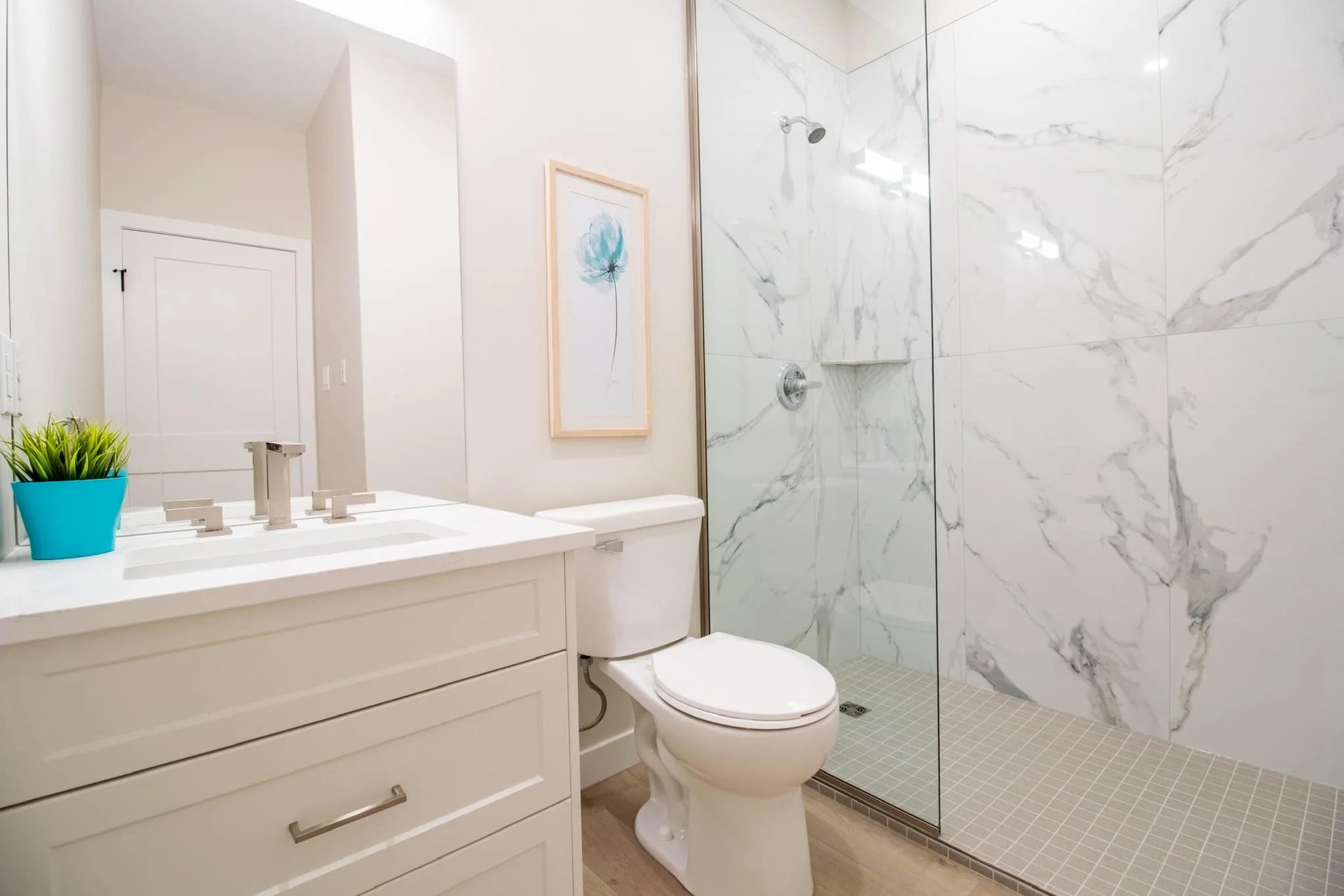 Modern bathroom with white vanity, potted green plant, framed art, white toilet, and glass-enclosed shower with marble wall tiles and beige floor tiles.