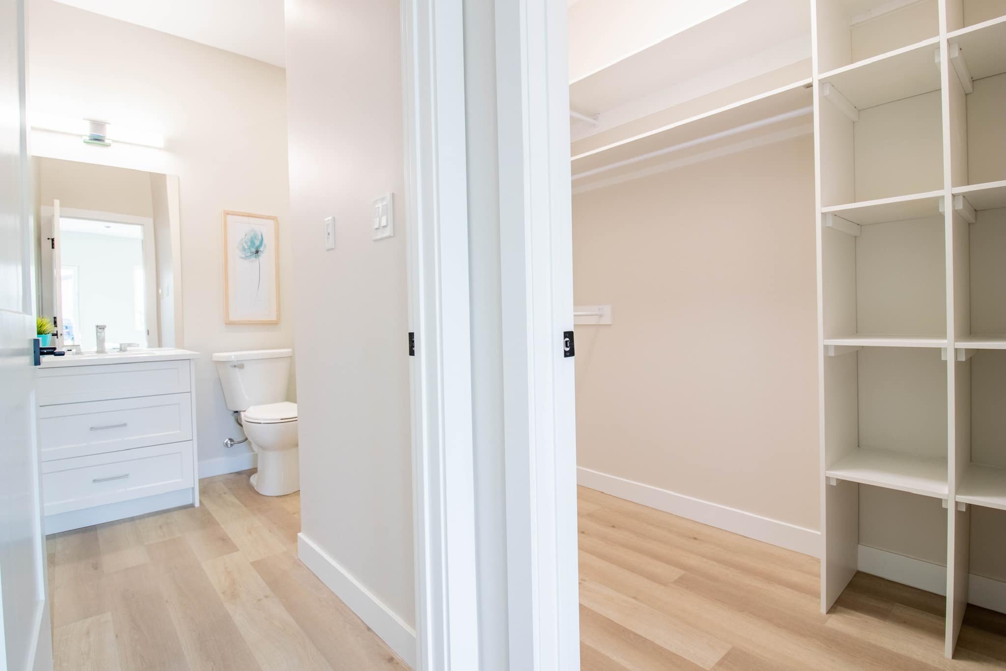 Interior view of a bathroom and an empty walk-in closet with white shelving, hardwood floors, and light-colored walls.