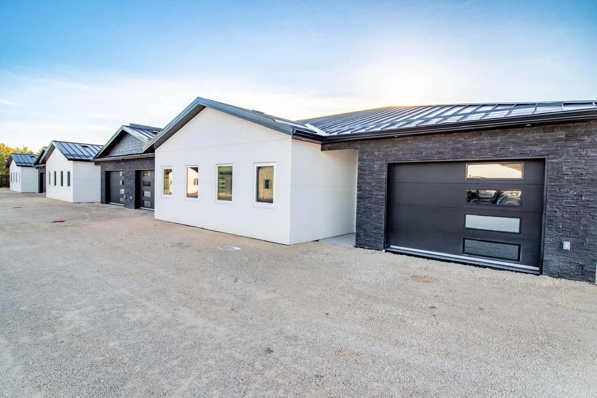 Row of modern houses with different exterior finishes, metal roofs, and black garage doors, set against a clear sky.