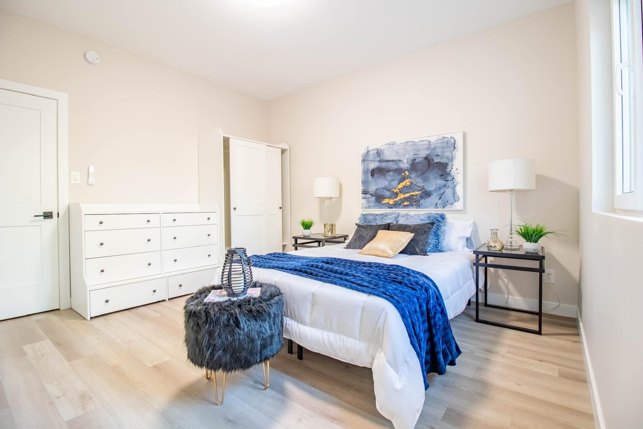 A modern bedroom with a white bed, blue throw blanket, beige and gray pillows, black side tables with lamps, potted plants, and abstract artwork on the wall. Light wood flooring and a window on the right side.