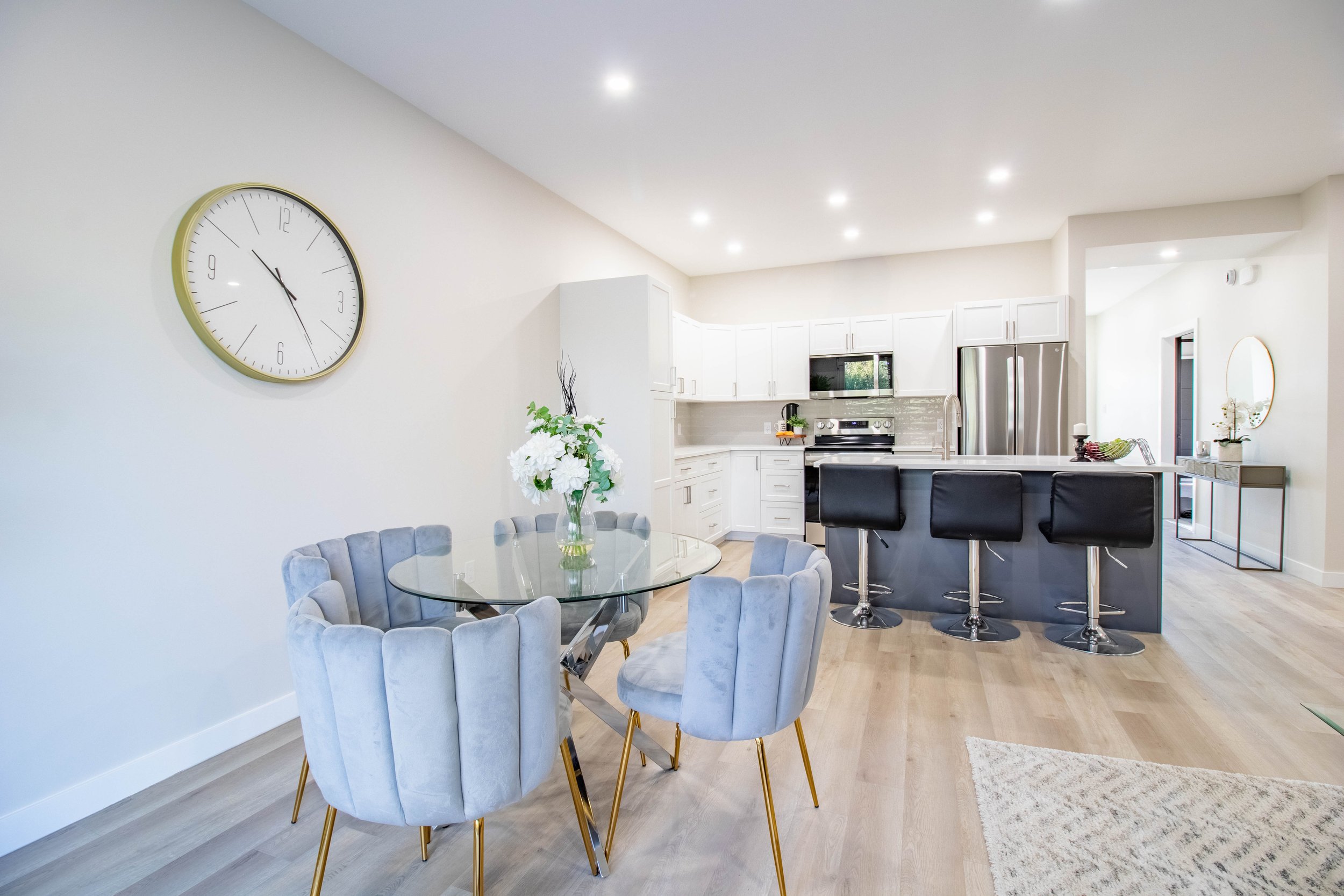 Open-concept kitchen and dining area with white cabinets, stainless steel appliances, a black kitchen island with four black bar stools, a round glass dining table with six gray upholstered chairs, and a large wall clock. Light wood flooring and minimal decor.
