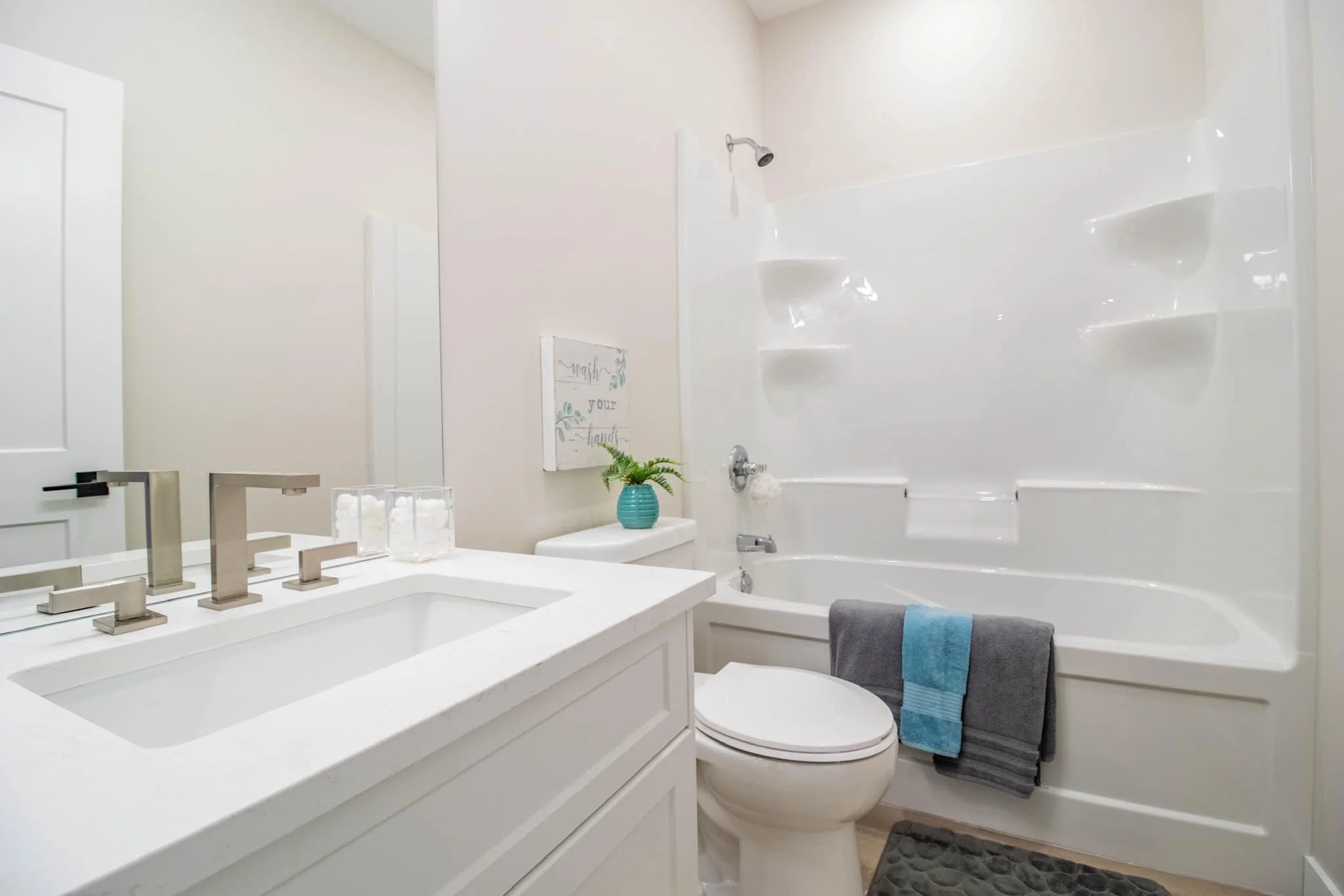 A clean bathroom with a white vanity and sink, a white toilet with a gray and blue towel hanging nearby, a white bathtub with built-in shelves, a small potted plant on the toilet tank, and a wall decor piece that says "Wash your hands".