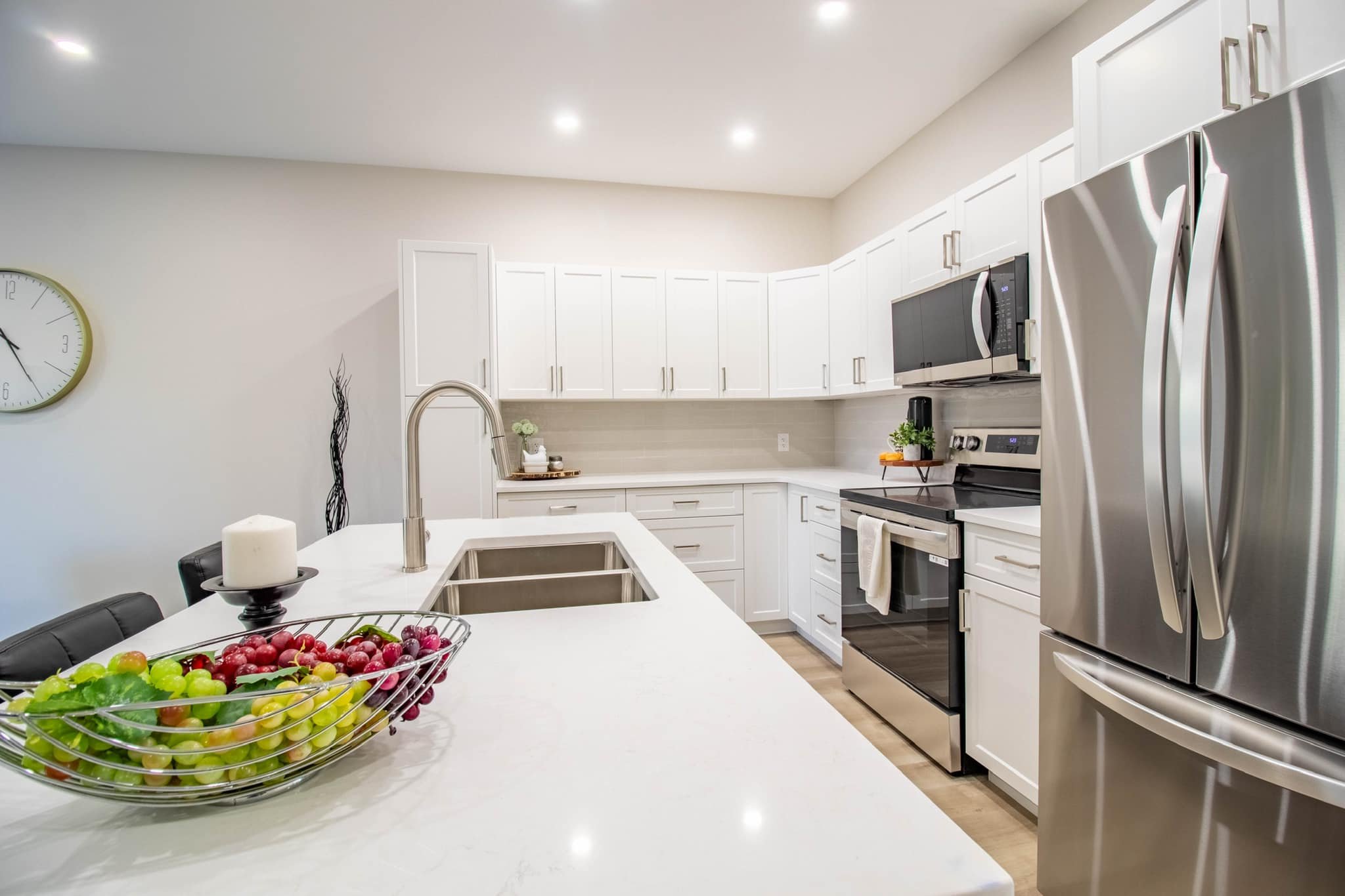 Modern white kitchen with stainless steel refrigerator, black stove and microwave, white cabinets, and a kitchen island with a bowl of grapes and apples.