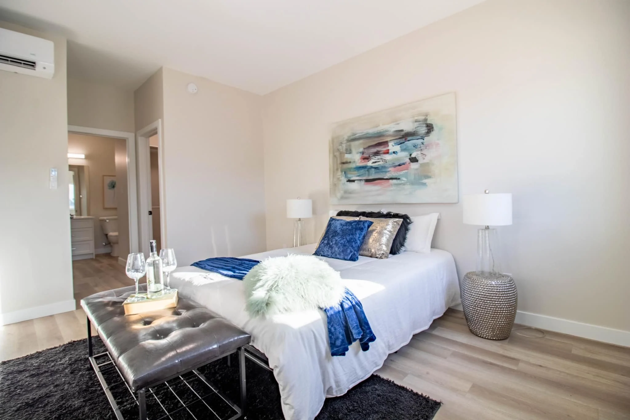 Modern bedroom with white bed, colorful abstract artwork above, side tables with lamps, black leather bench with drinks, and sliding door to bathroom