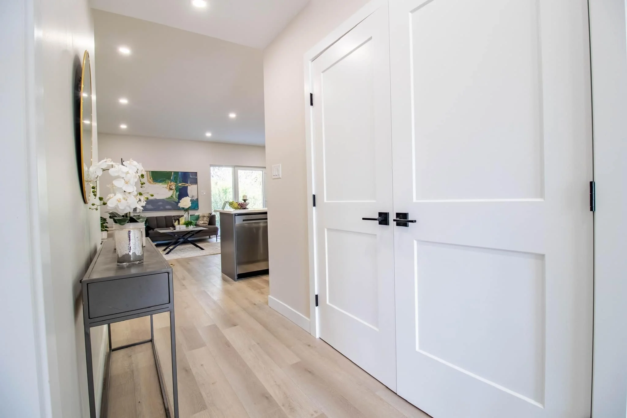 Entrance hallway leading into a living room with a sofa, artwork on the wall, and large windows. The hallway has a gray console table with white flowers and a mirrored wall decor, and a set of white double doors to the right.